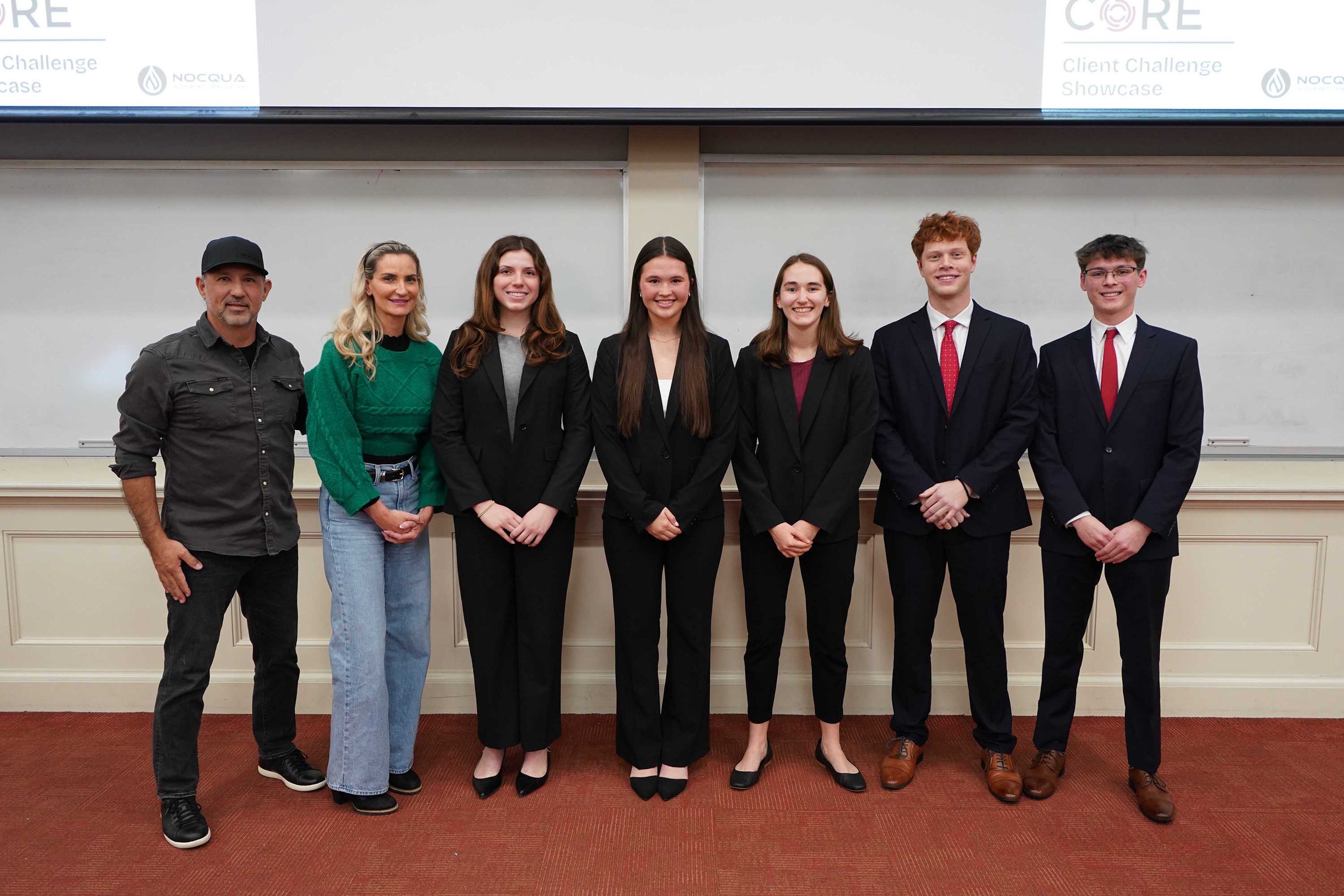Group photo of six individuals dressed in business attire, standing in front of a presentation backdrop during a client challenge showcase event.