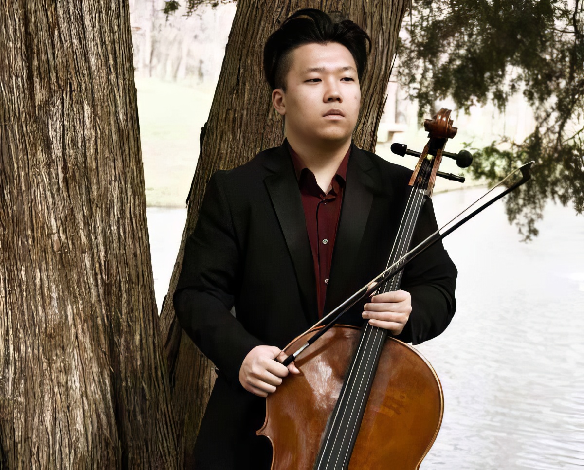 Marcus Cheung in formal attire stands by a tree near a body of water, holding his cello with a focused expression.