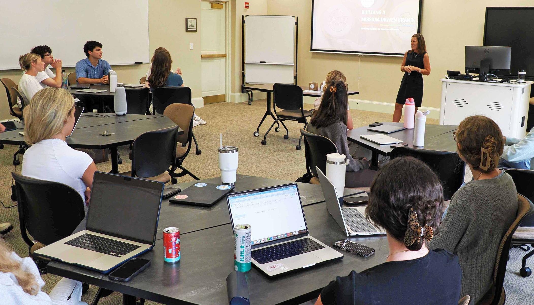 A group of students attentively listening to a presentation in a classroom setting, with laptops and drinks on the tables.