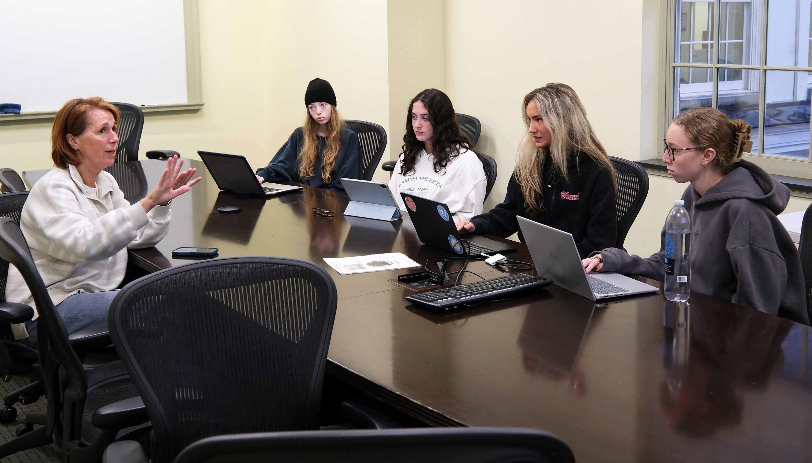A group of five young women engaged in a discussion around a conference table with laptops open in front of them.