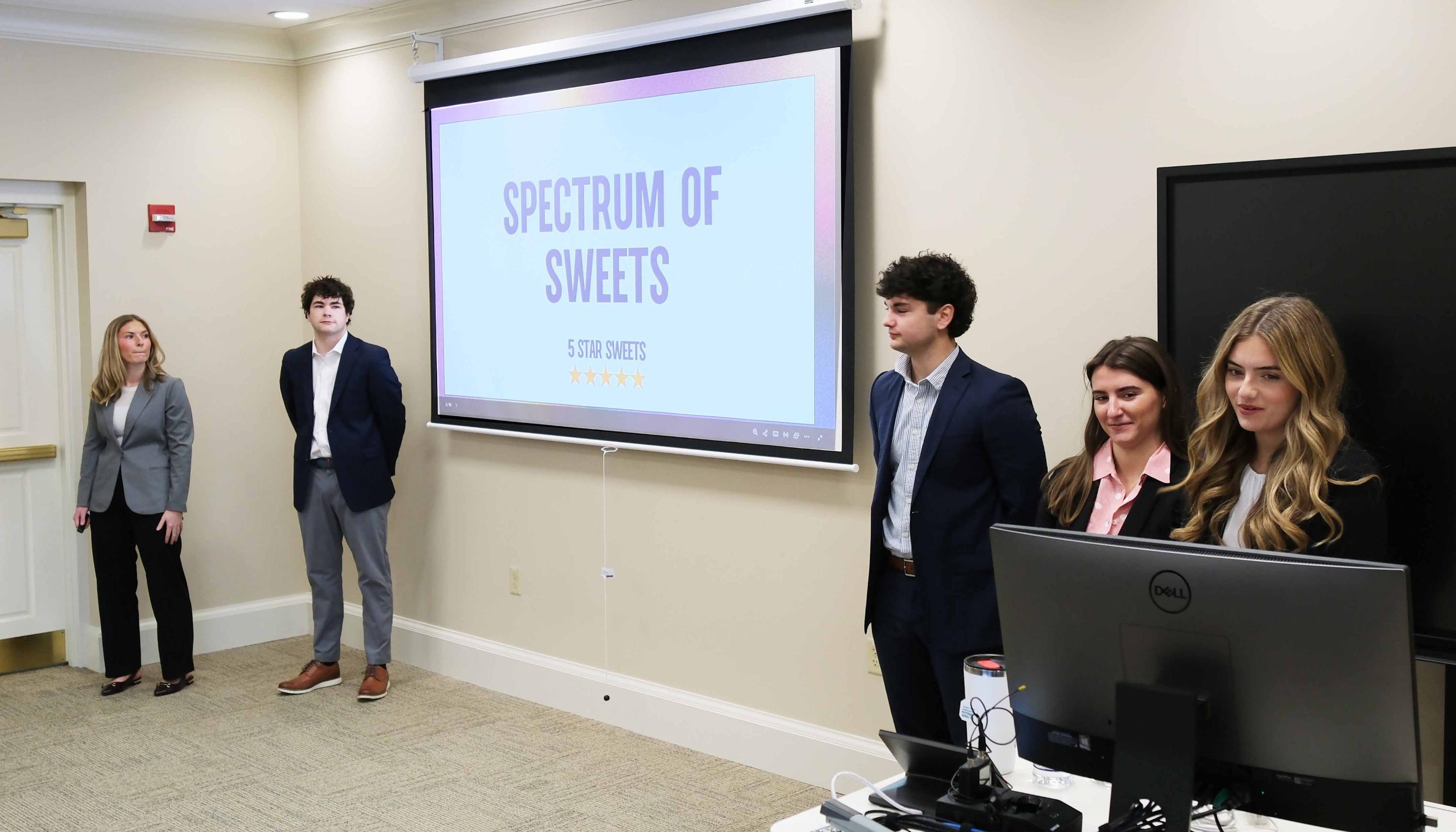 A team of four young professionals presents a project titled "Spectrum of Sweets" in a conference room, with a projector displaying the title on a screen.