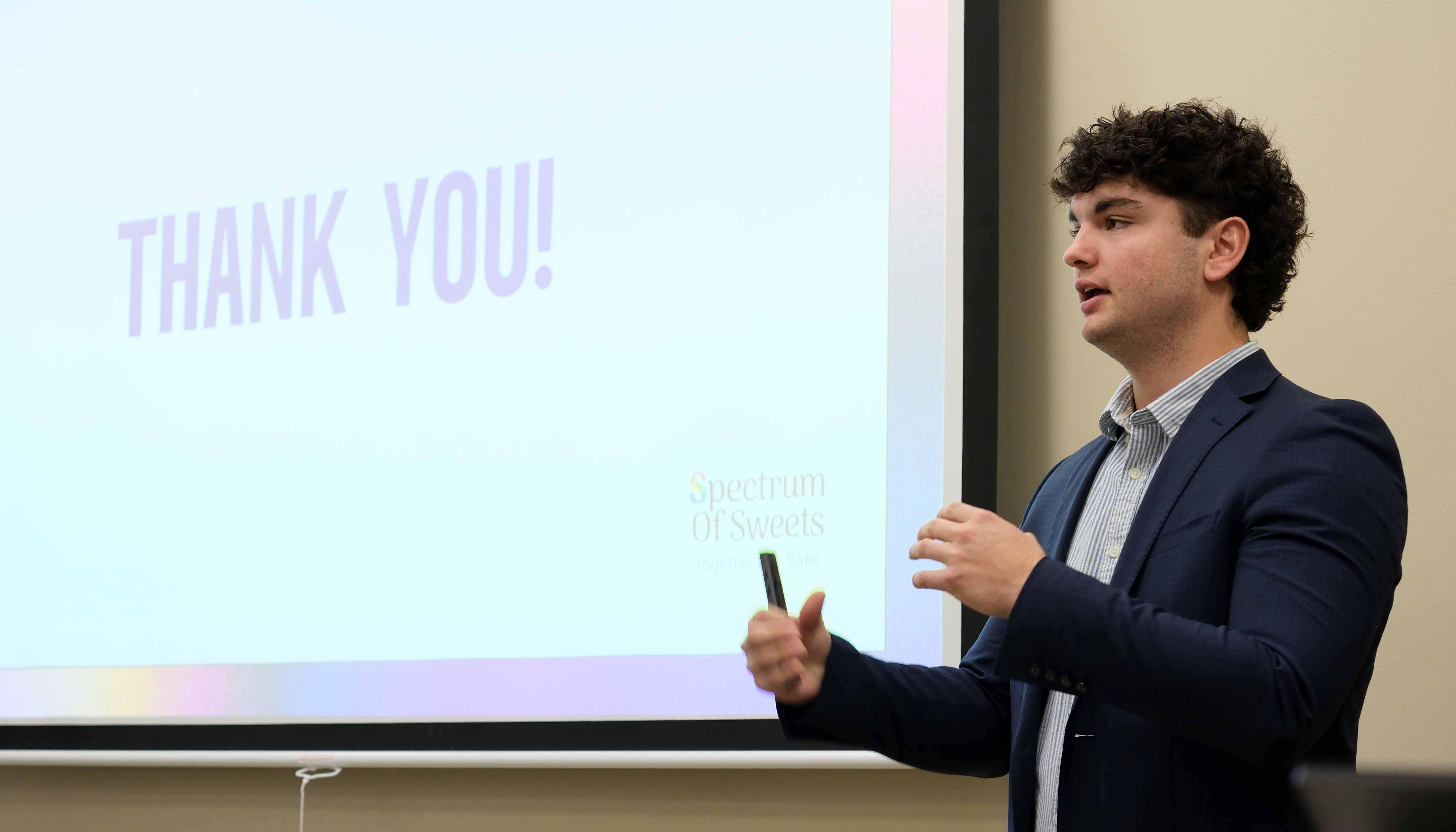 A young man in a blazer gestures while giving a presentation, with "THANK YOU!" displayed on a screen behind him.