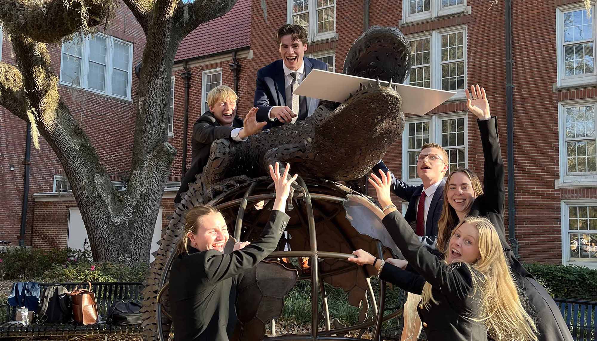 Group of students in formal attire posing excitedly on a large bronze alligator sculpture in front of a brick campus building.