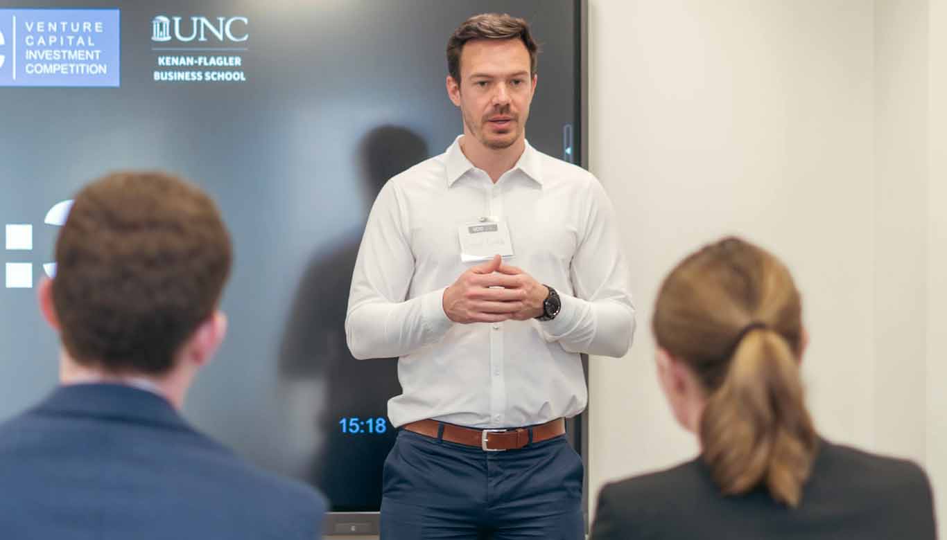Presenter speaking to an audience in front of a screen displaying UNC Kenan-Flagler Business School and Venture Capital Investment Competition branding.