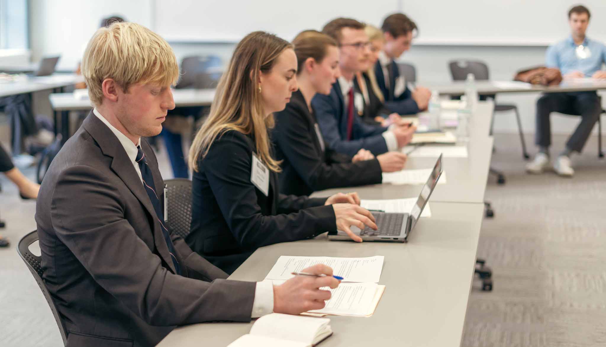 Students in business attire sit at a conference table taking notes and working on laptops during a classroom session.