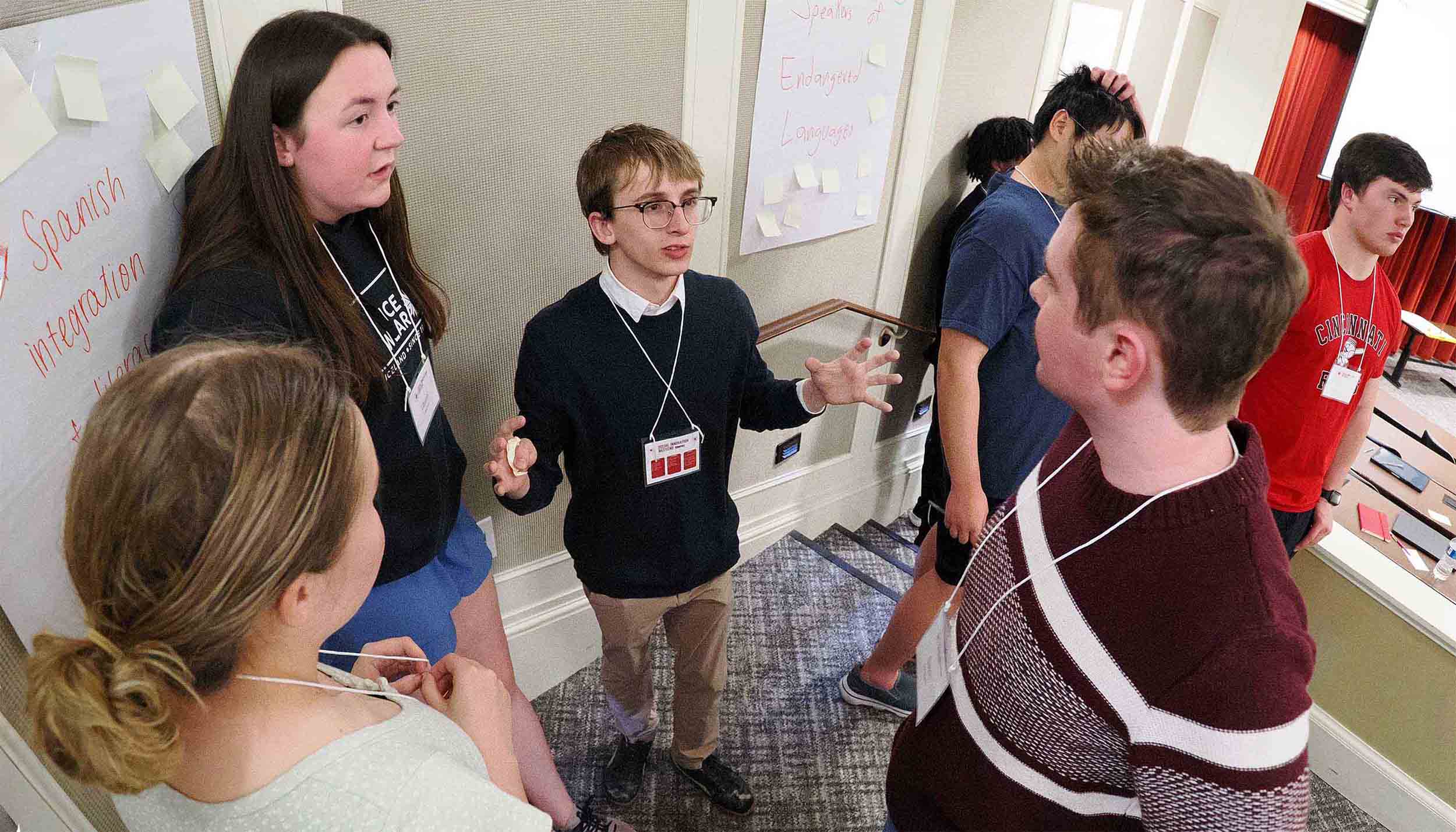 Students wearing name badges discuss ideas during a workshop session in a conference hallway.