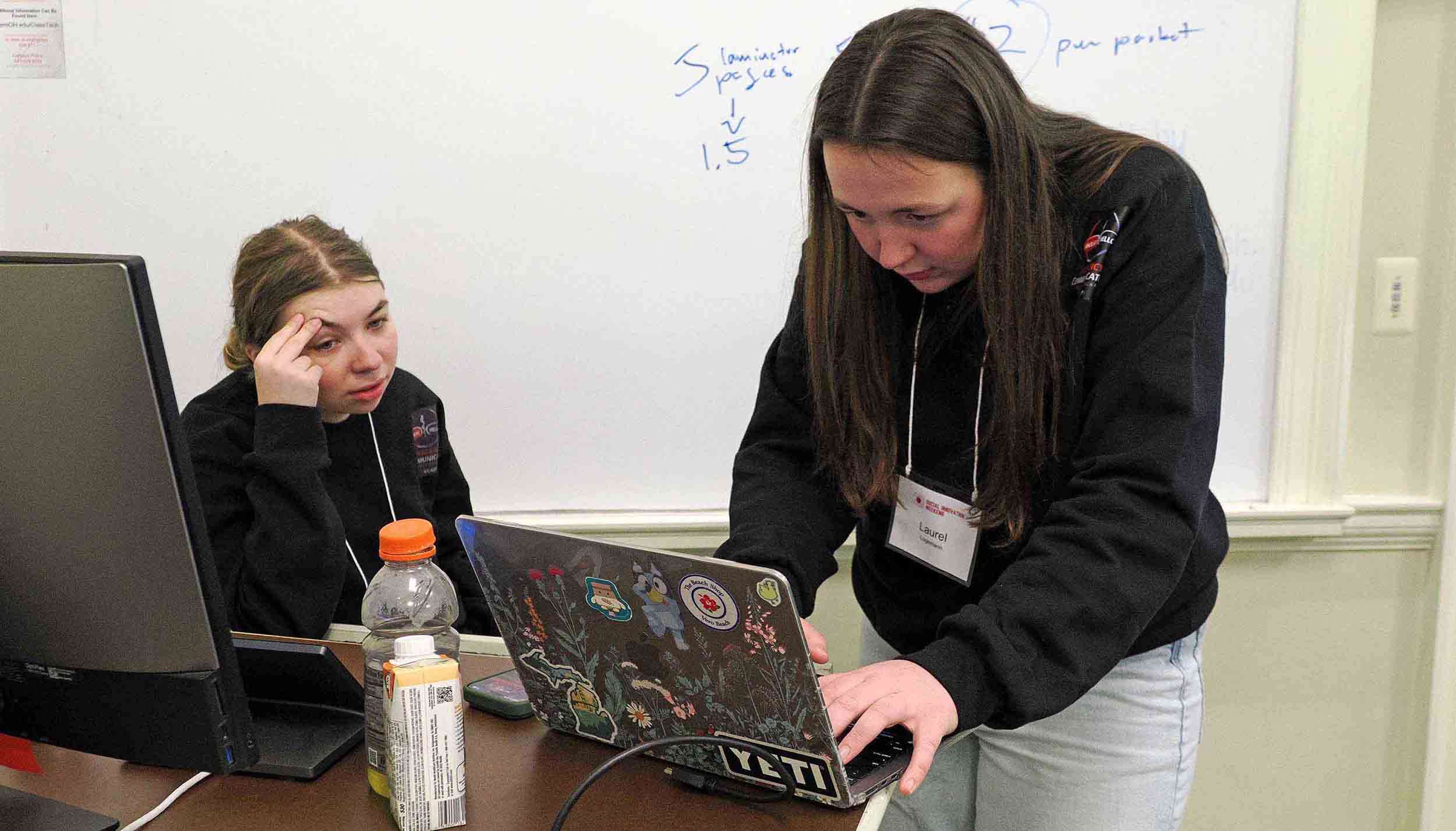 Two students work on a laptop together in a classroom, with a whiteboard in the background.
