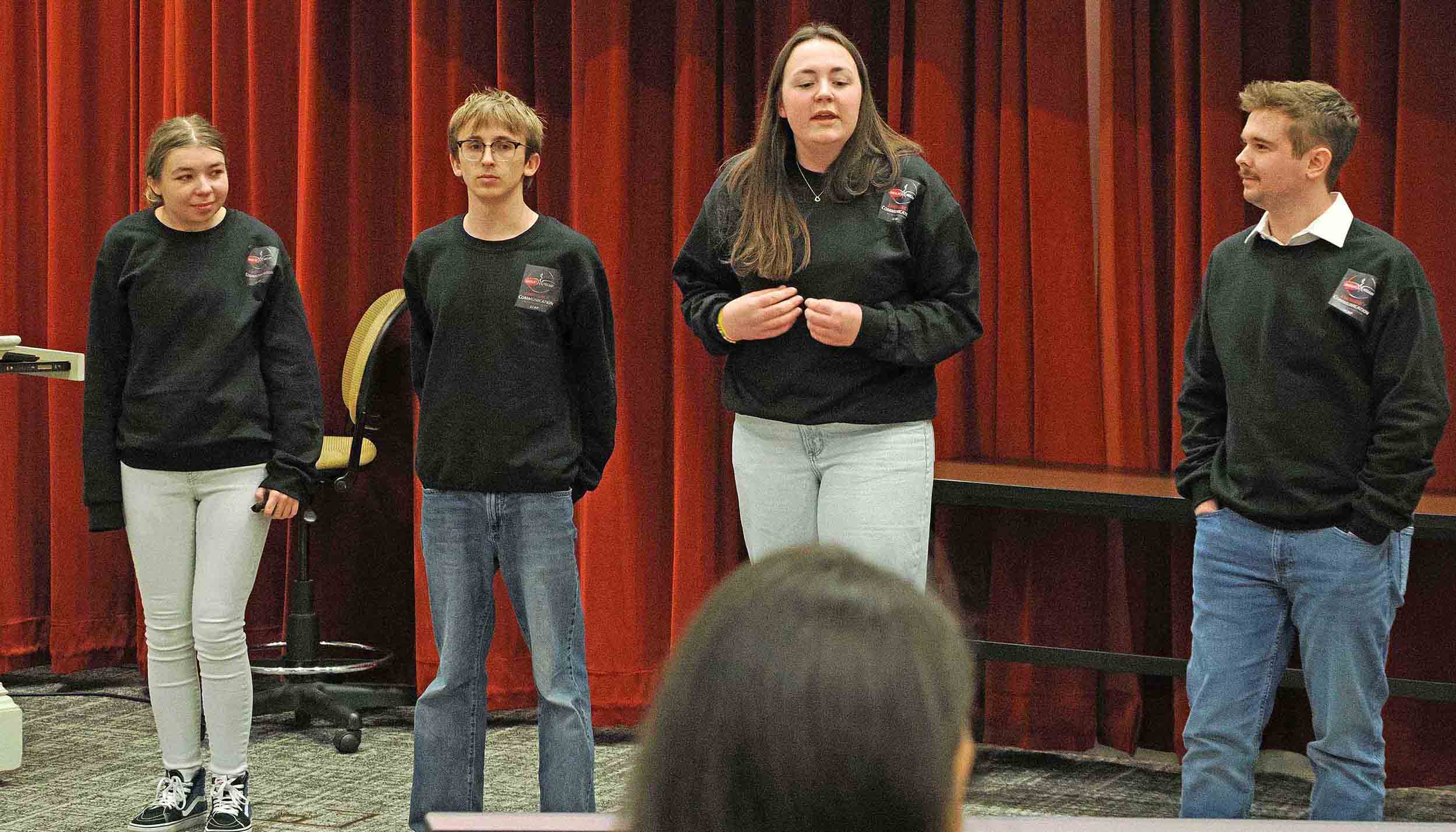 Four presenters in matching black sweatshirts stand in front of red curtains, speaking to an audience.