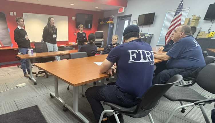 Fire department meeting in a training room, with several firefighters seated at tables while three people speak at the front.