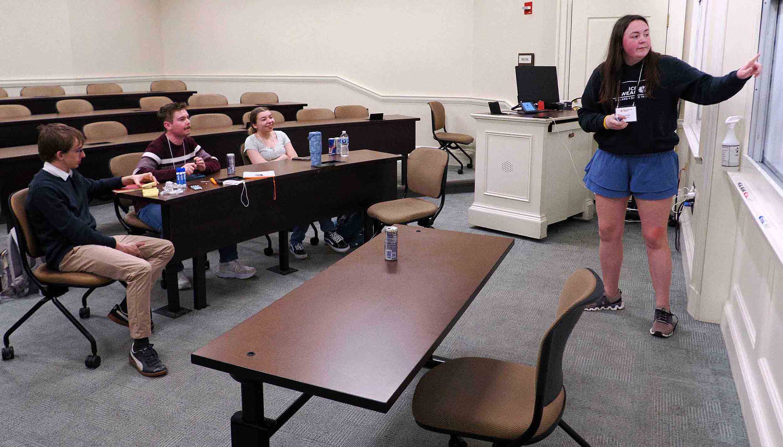 Student presenter points to a whiteboard while classmates listen in a lecture classroom.