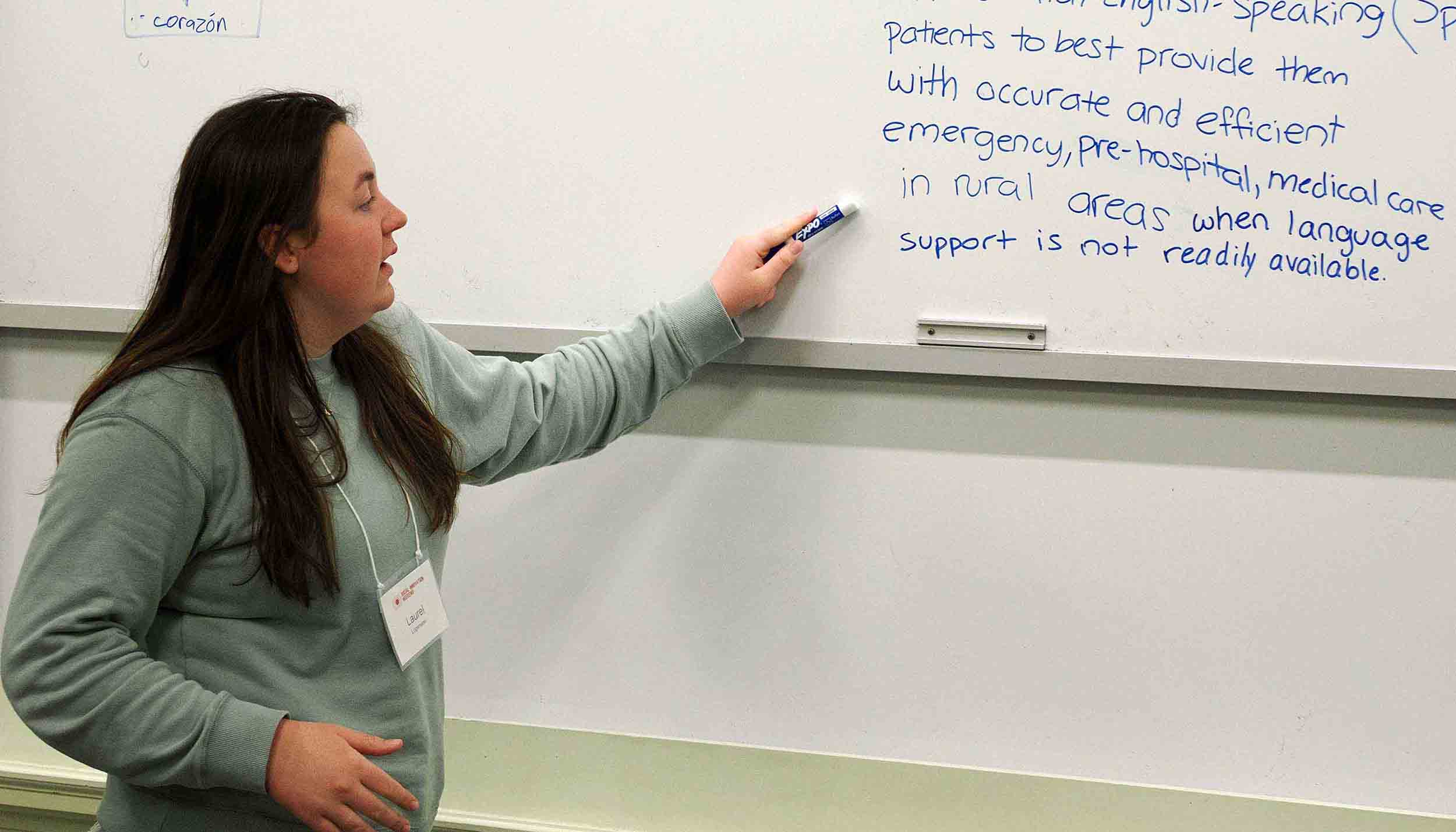 Person presenting in a classroom, pointing at notes written on a whiteboard.
