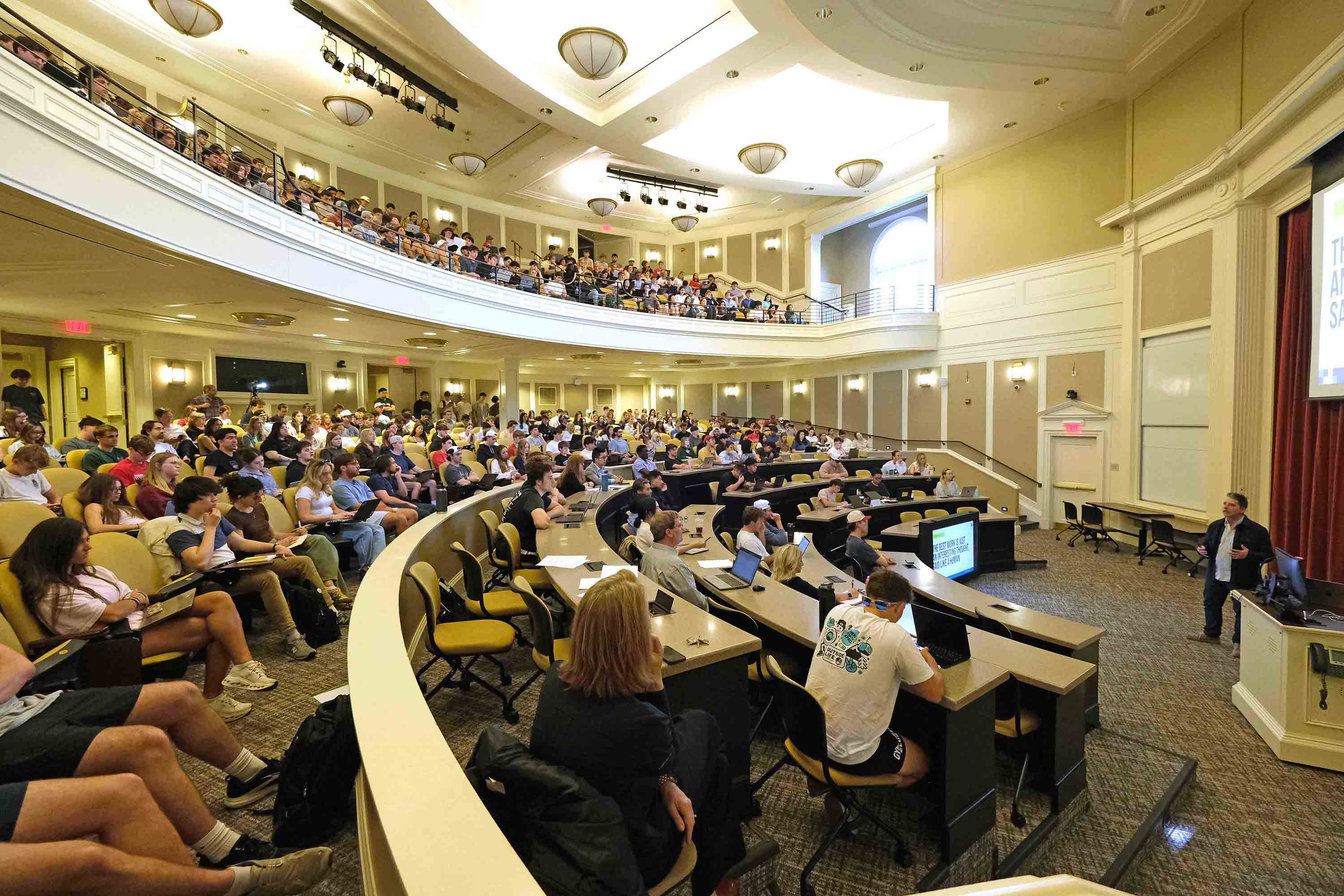 Packed university lecture hall with students seated in tiered rows while an instructor presents at the front.