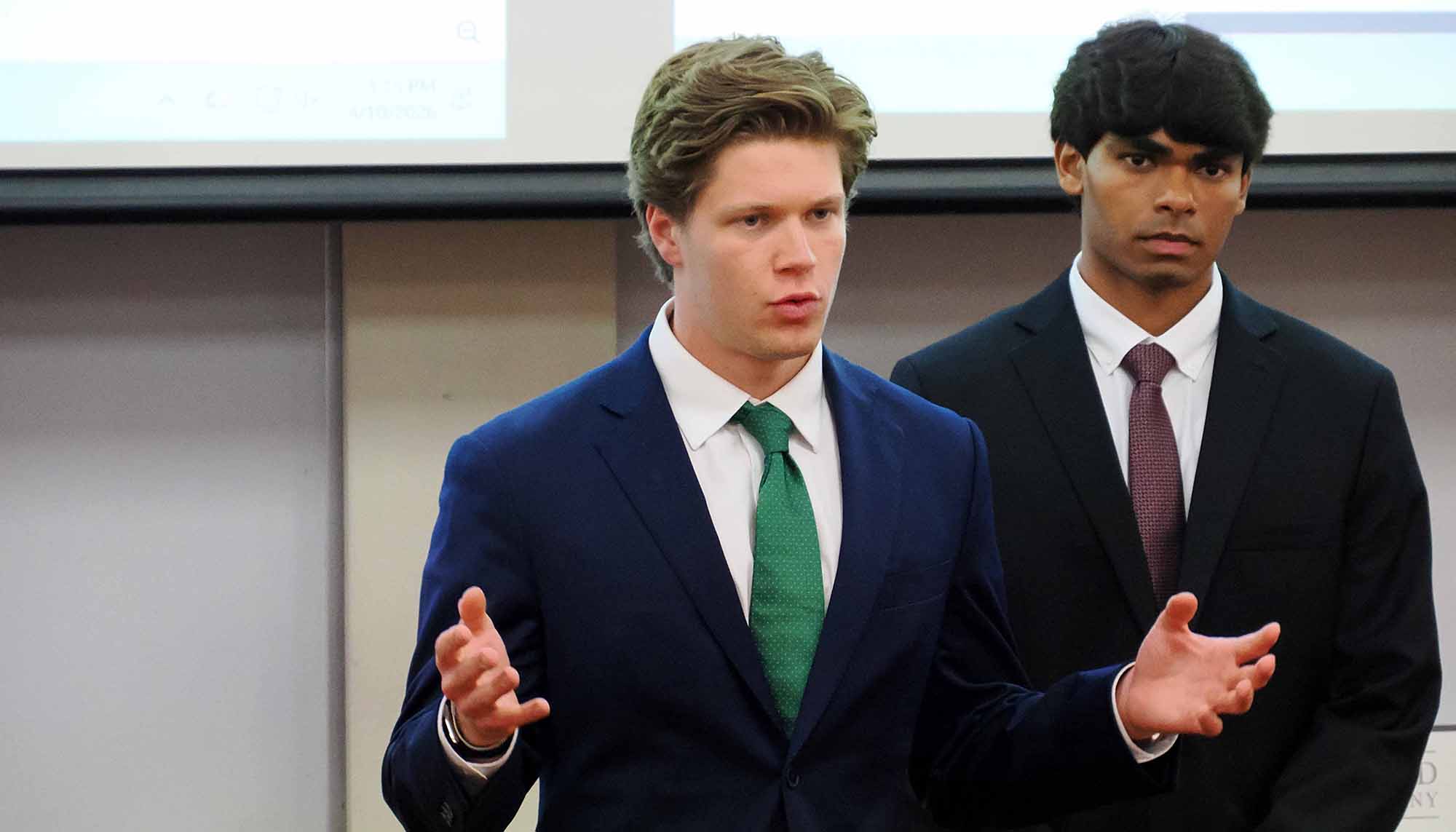 Two men in suits give a presentation in front of a projection screen, with one speaking and gesturing while the other stands beside him.
