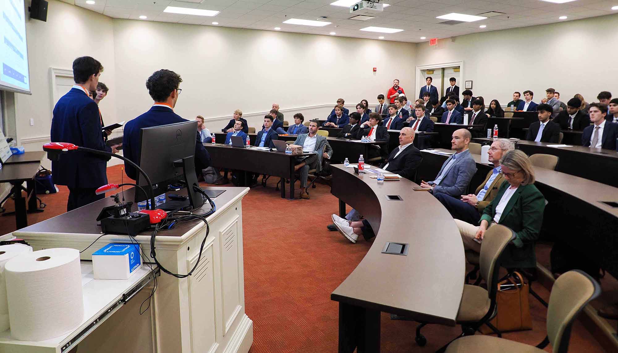 Two presenters in suits address a seated audience in a lecture hall during a formal presentation.