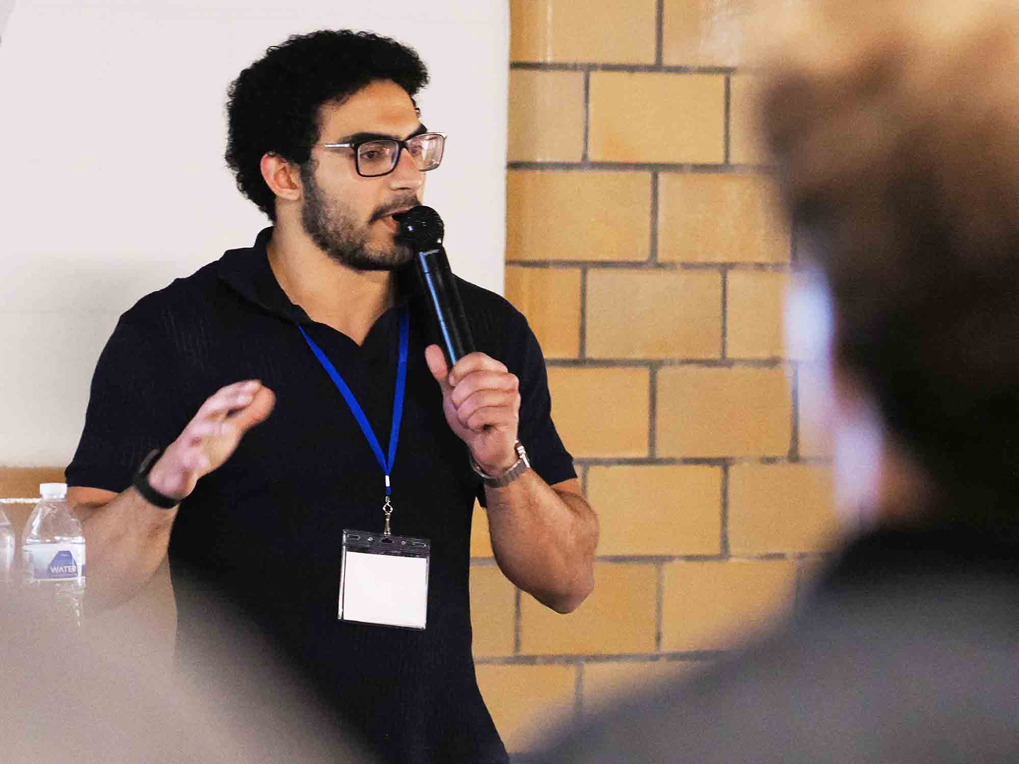 Speaker wearing a conference badge talks into a microphone while addressing an audience.