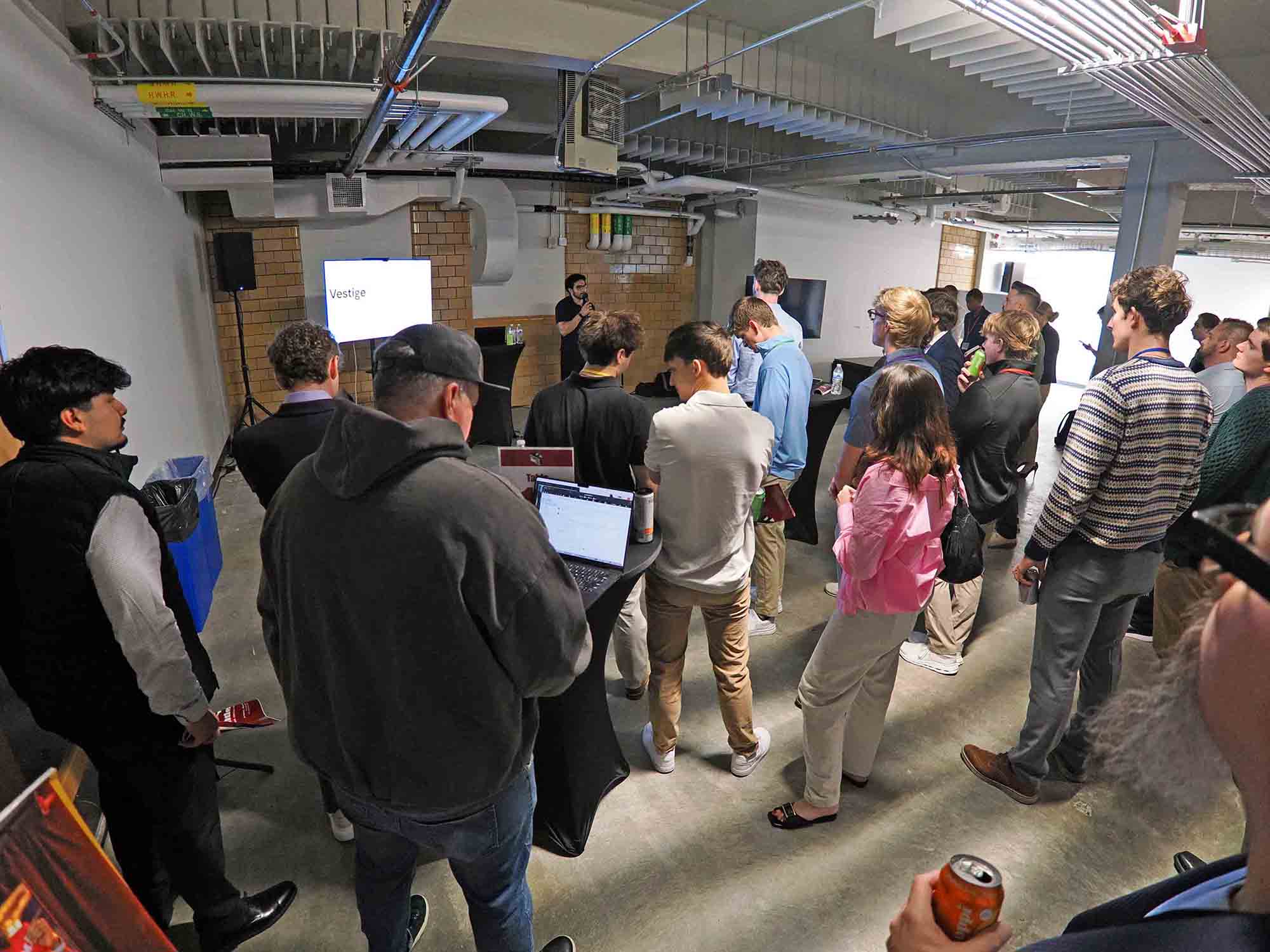 Audience gathered in an industrial-style room watching a presenter speak beside a projected slide.