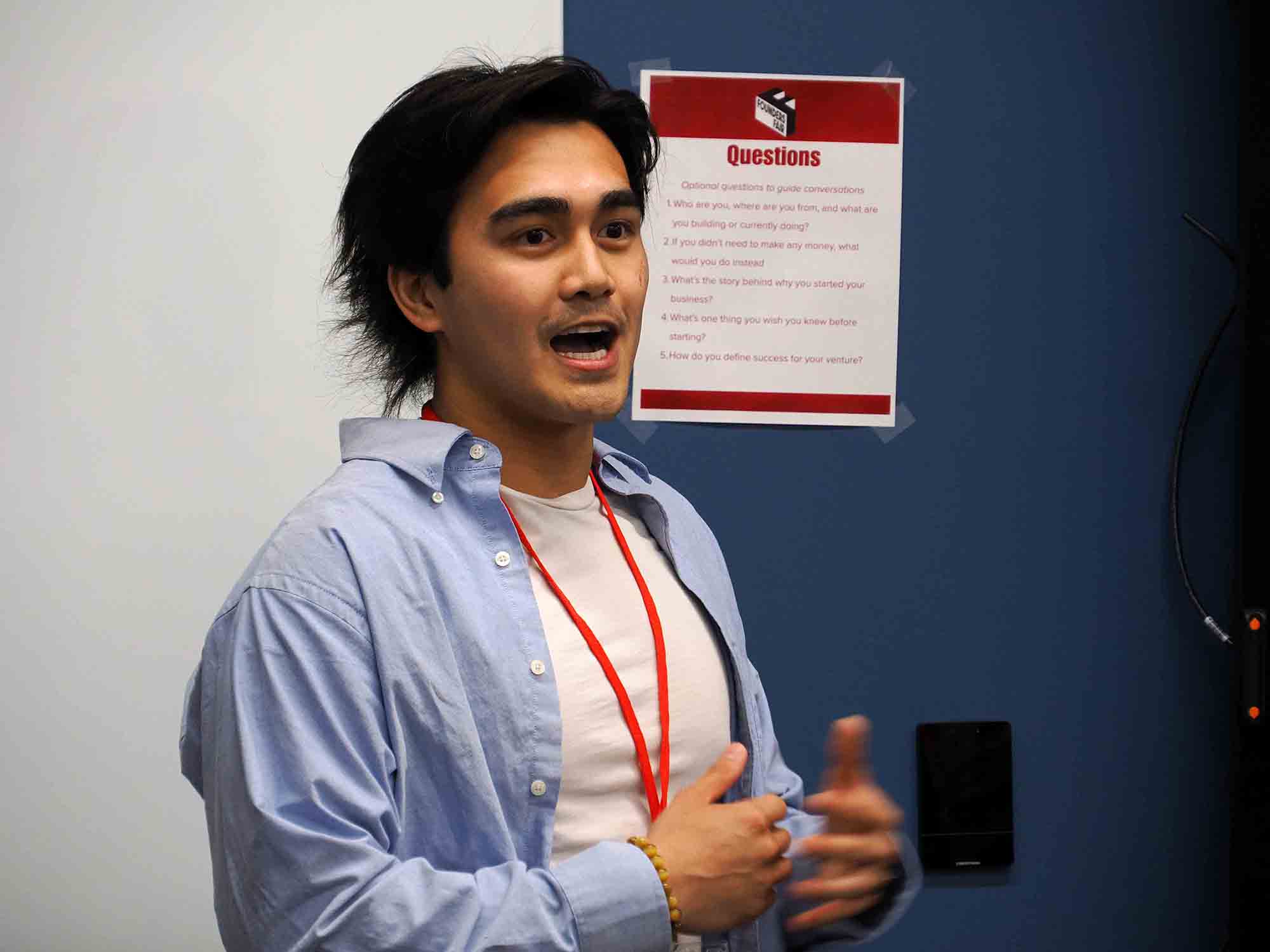 Person speaking and gesturing during a discussion, standing beside a wall poster titled “Questions.”