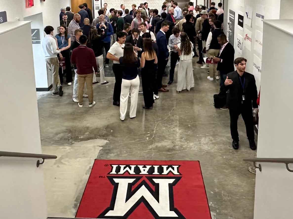 Crowd of attendees mingling in a hallway at an event, with a University of Miami “M” logo mat in the foreground.