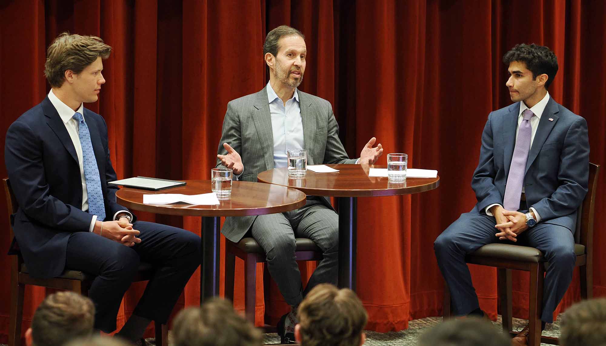 Three men in suits seated at small tables on a stage, having a panel discussion in front of red curtains.