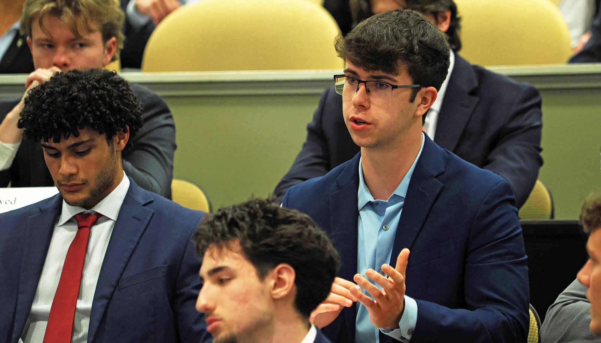 Young man in a suit and glasses speaks and gestures during a meeting while others listen in the background.