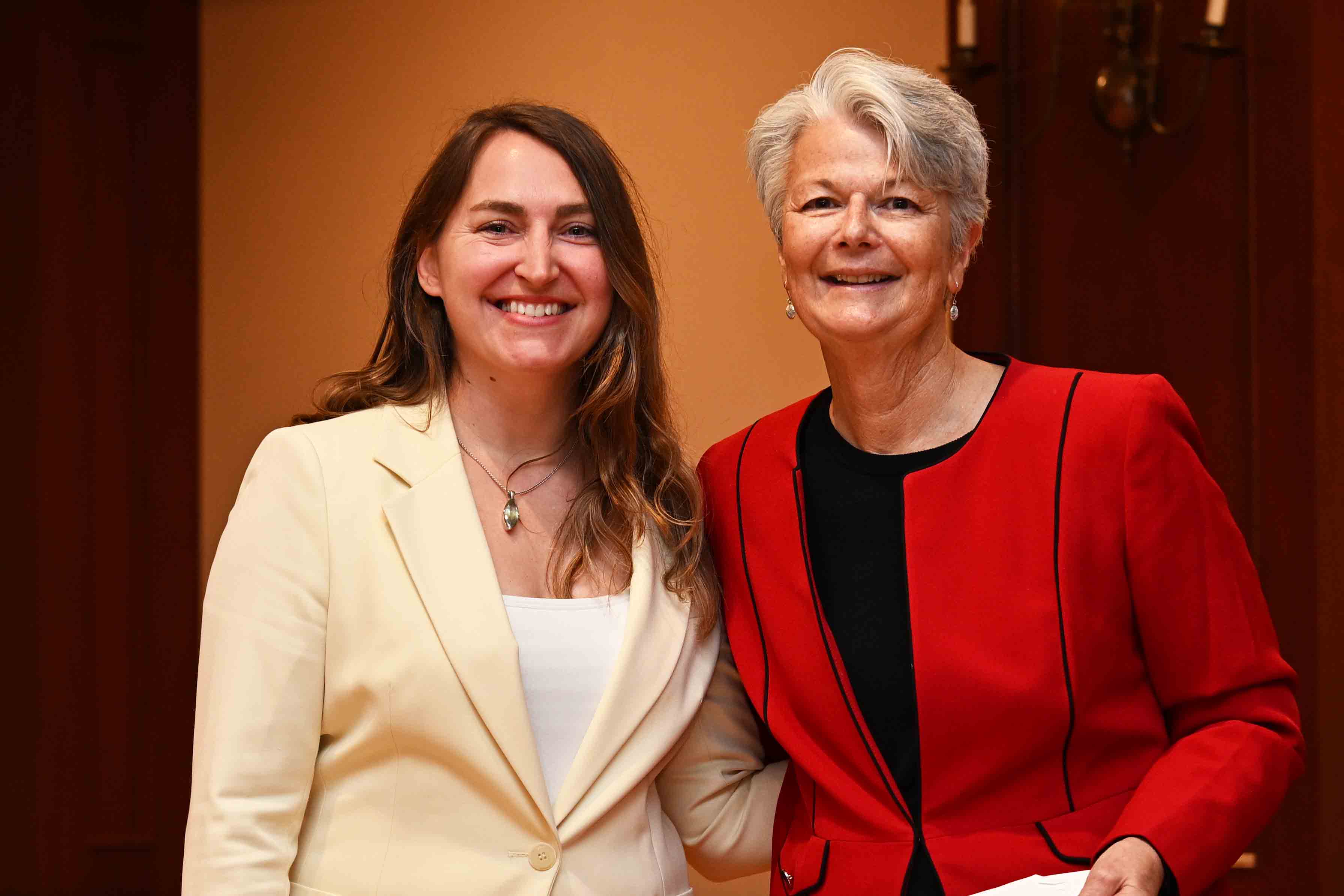 Two women smiling and posing together indoors, one in a cream blazer and the other in a red jacket.