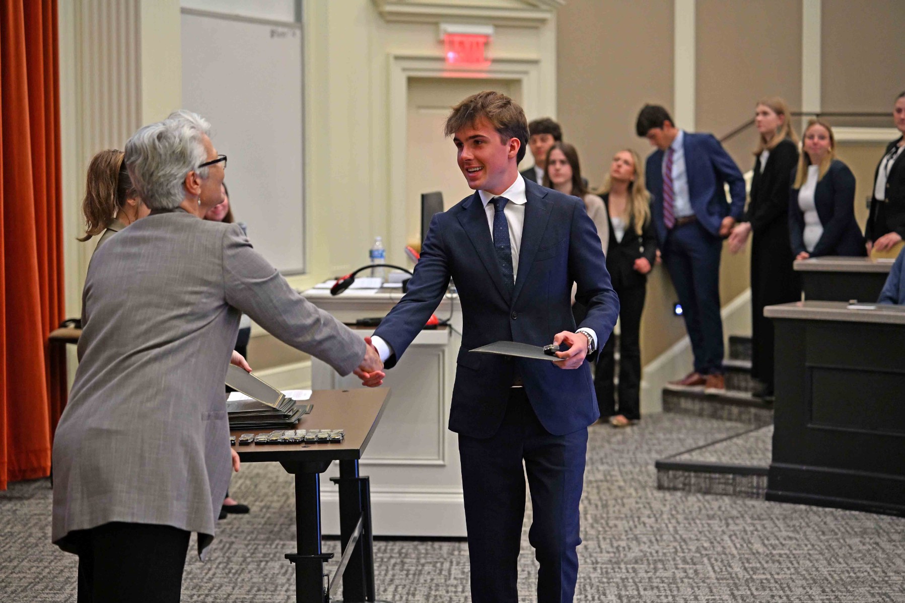 A student in a suit shakes hands with an organizer while receiving an award at a ceremony, with other students waiting in the background.