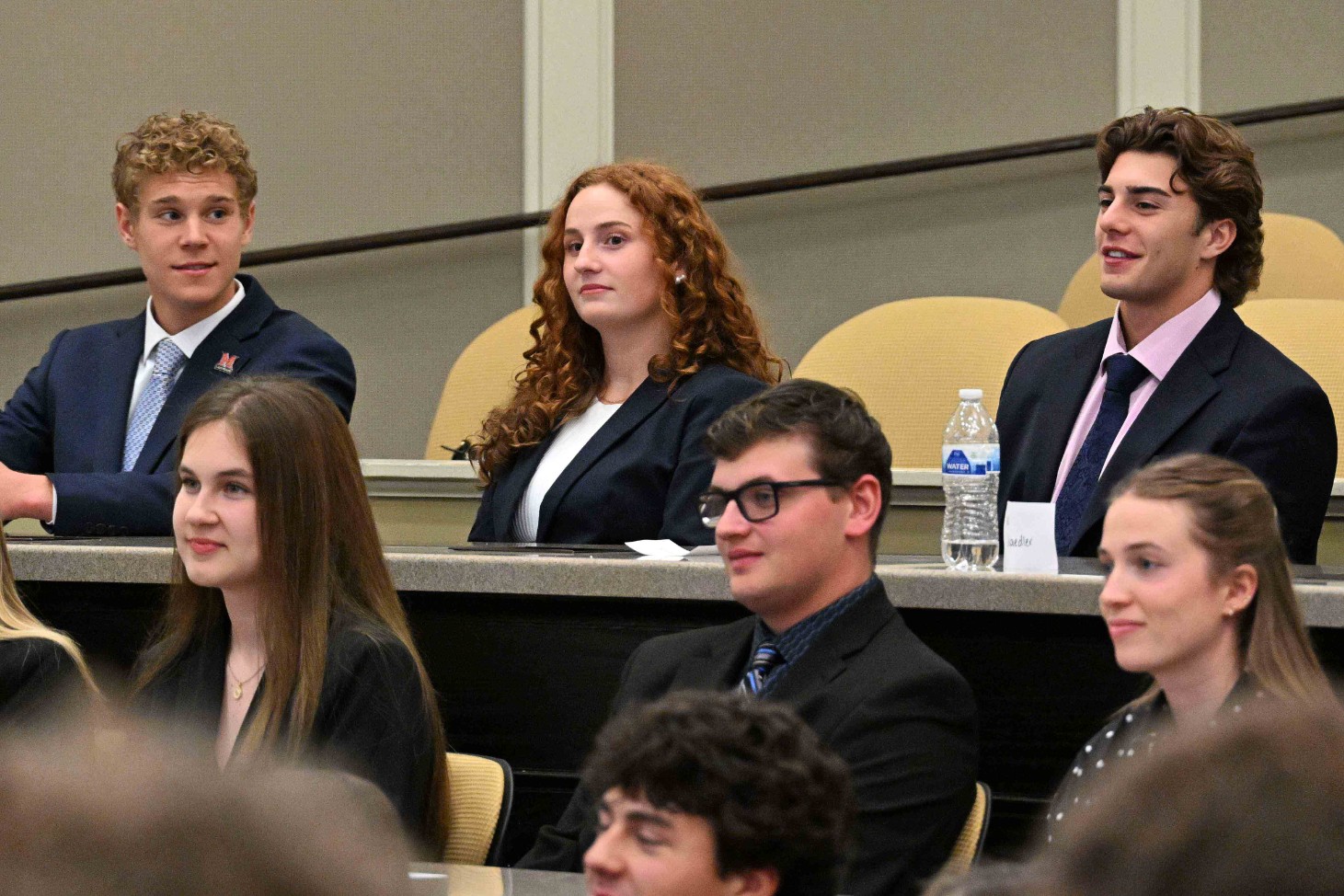 Students in business attire seated in a lecture hall during a formal event.