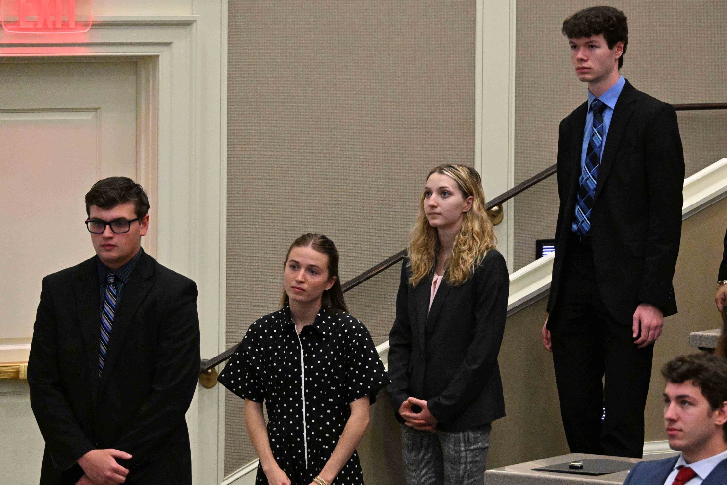 Four young adults in business attire standing indoors, as if at a formal ceremony or presentation.