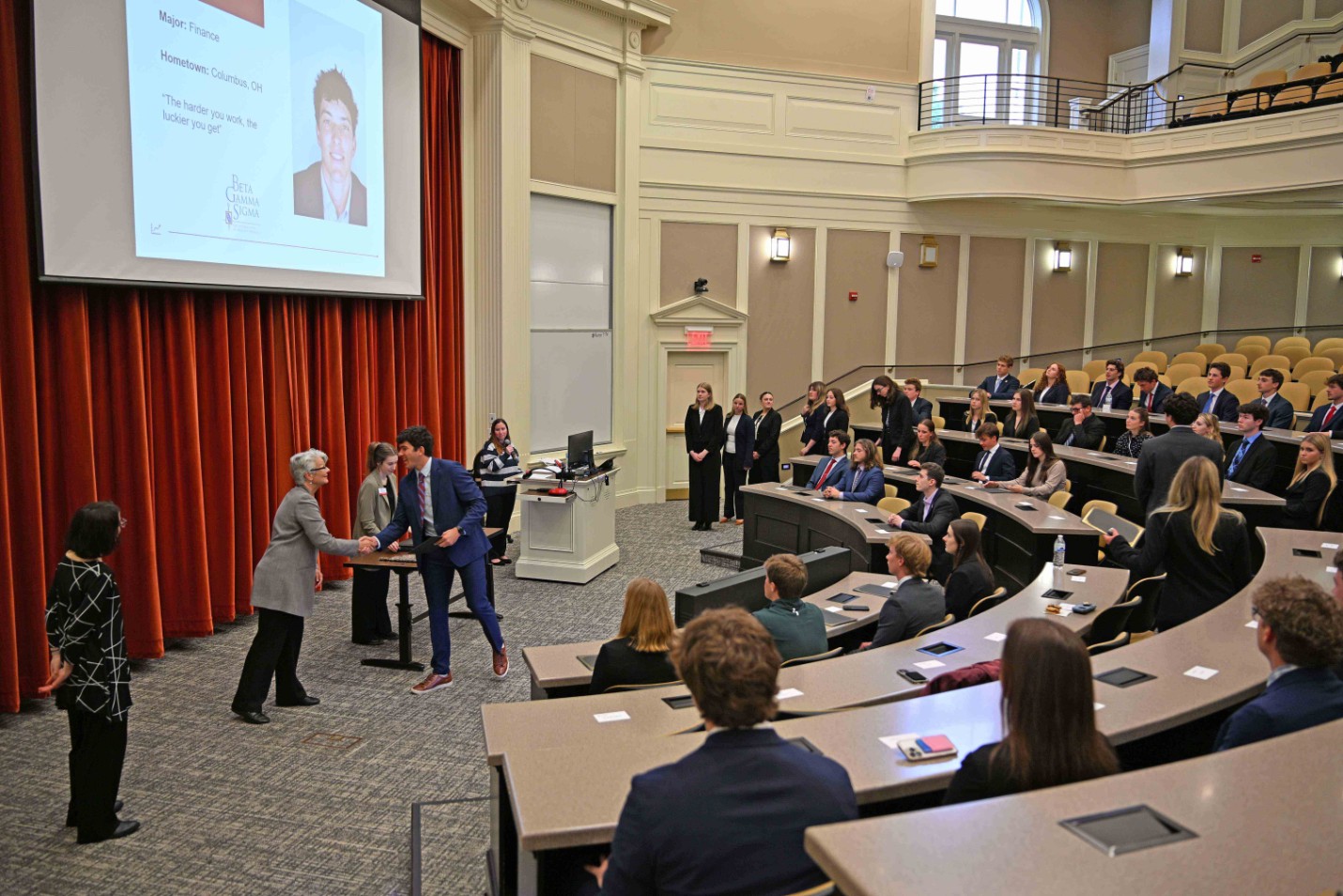 Speaker shaking hands with a student in a lecture hall while an audience in business attire watches.