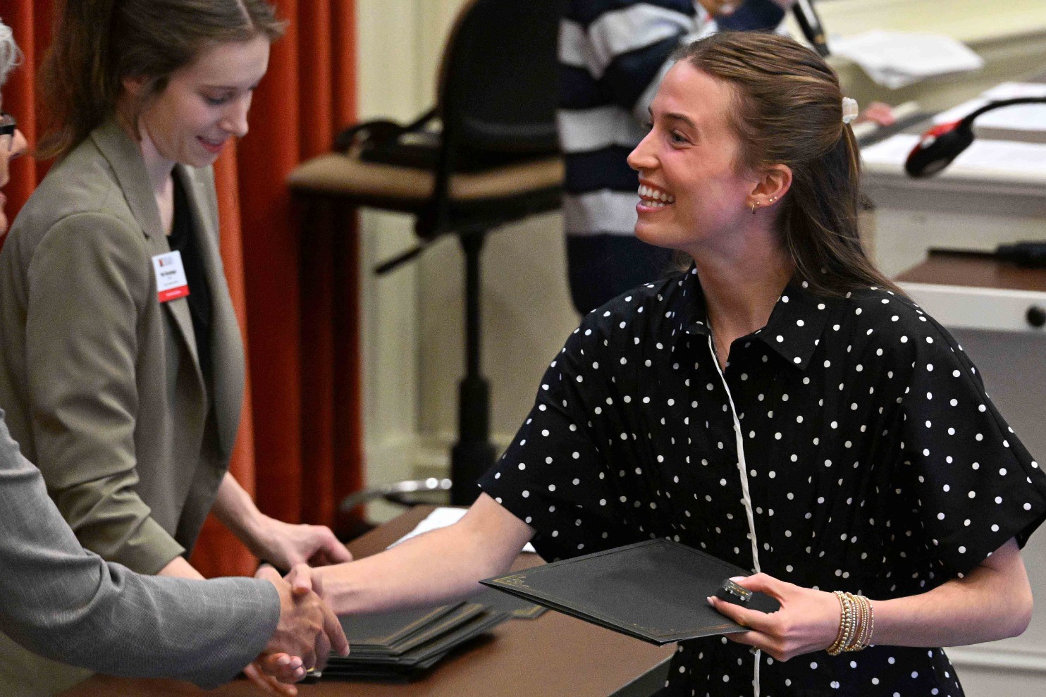 Smiling woman in a polka-dot blouse shakes hands while holding a certificate at an indoor awards event.
