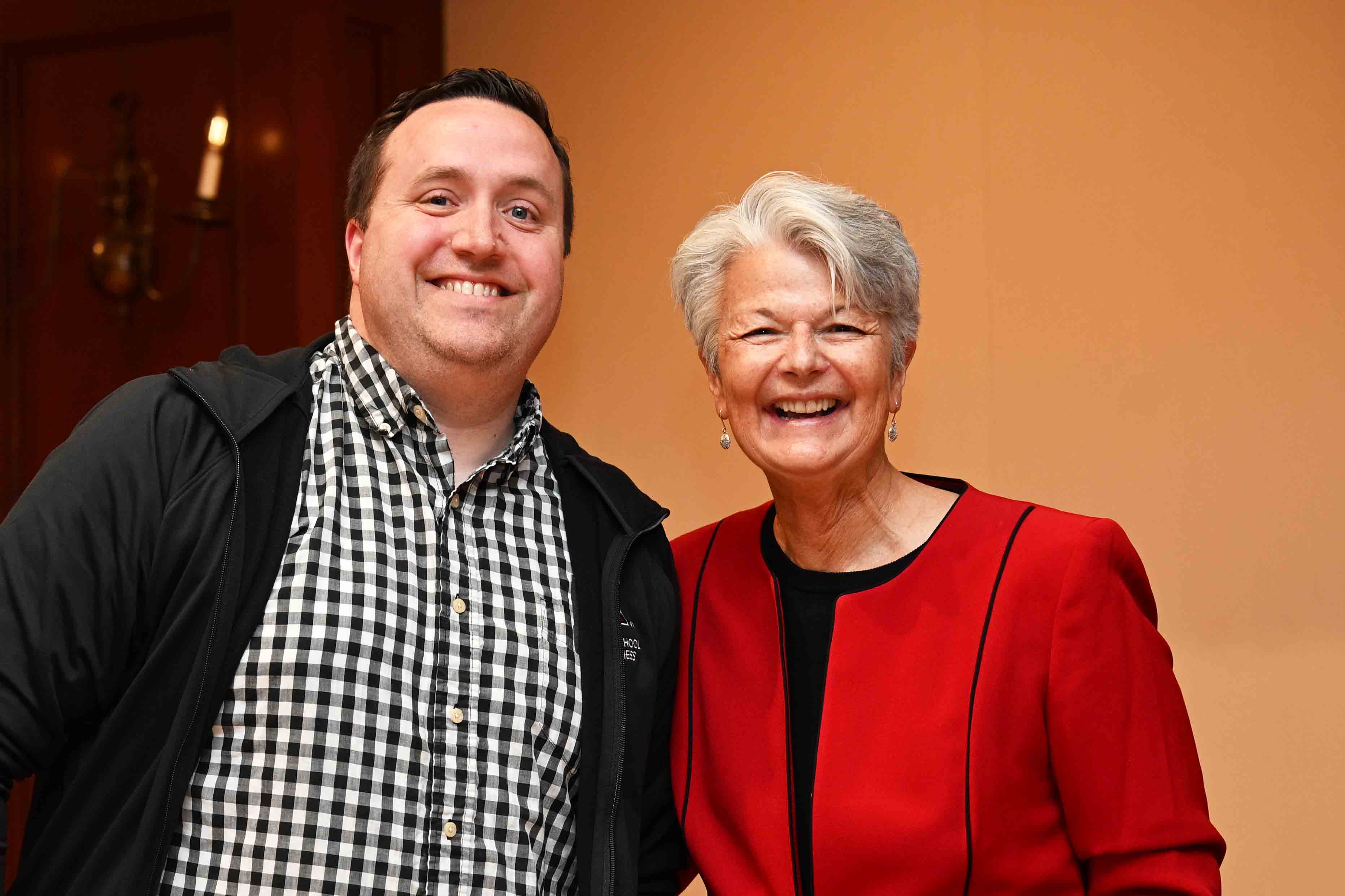 Two people smiling together indoors, one wearing a checkered shirt and jacket and the other in a red blazer.
