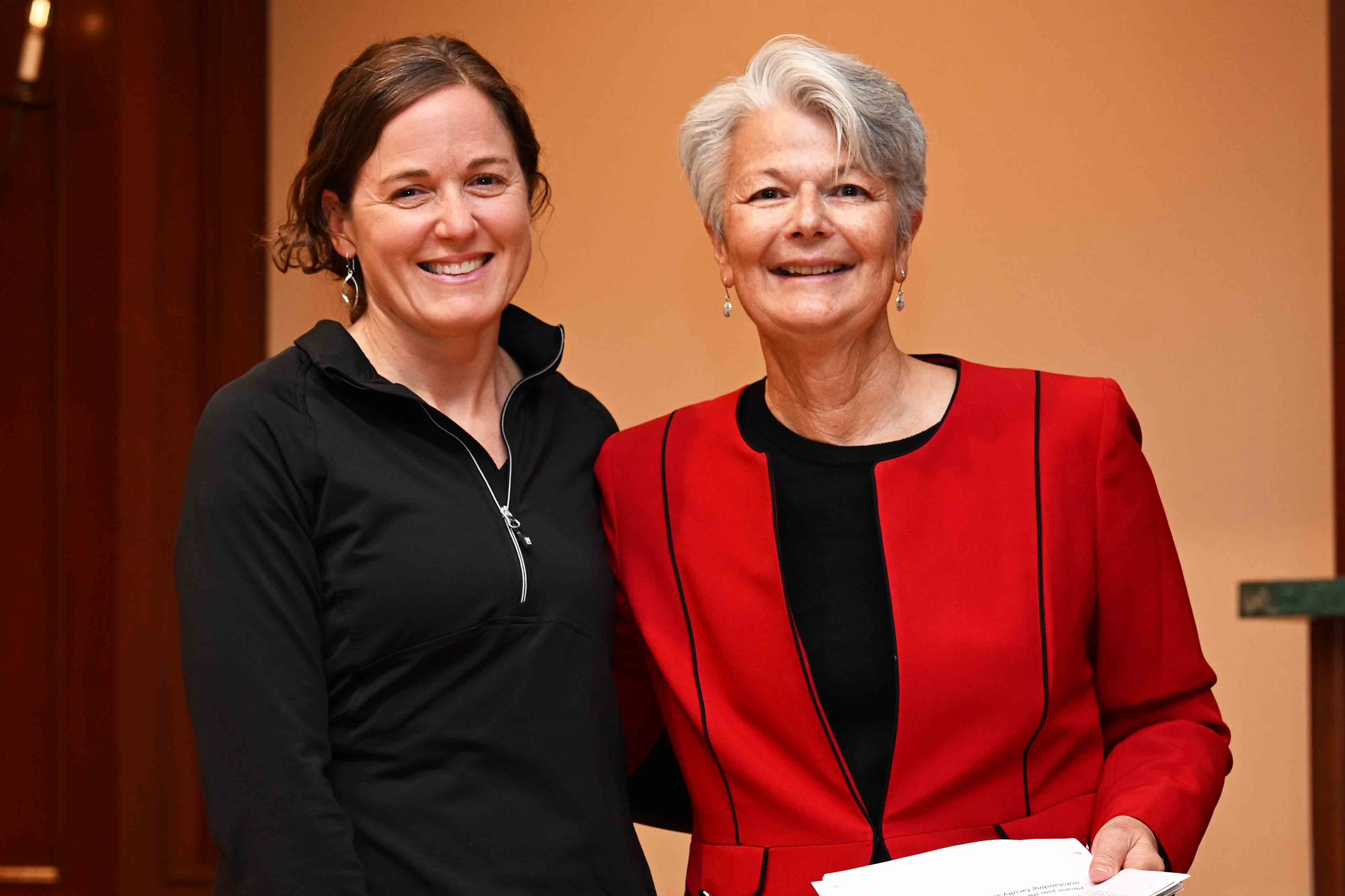 Two women smiling indoors, one in a black top and one in a red blazer holding papers.