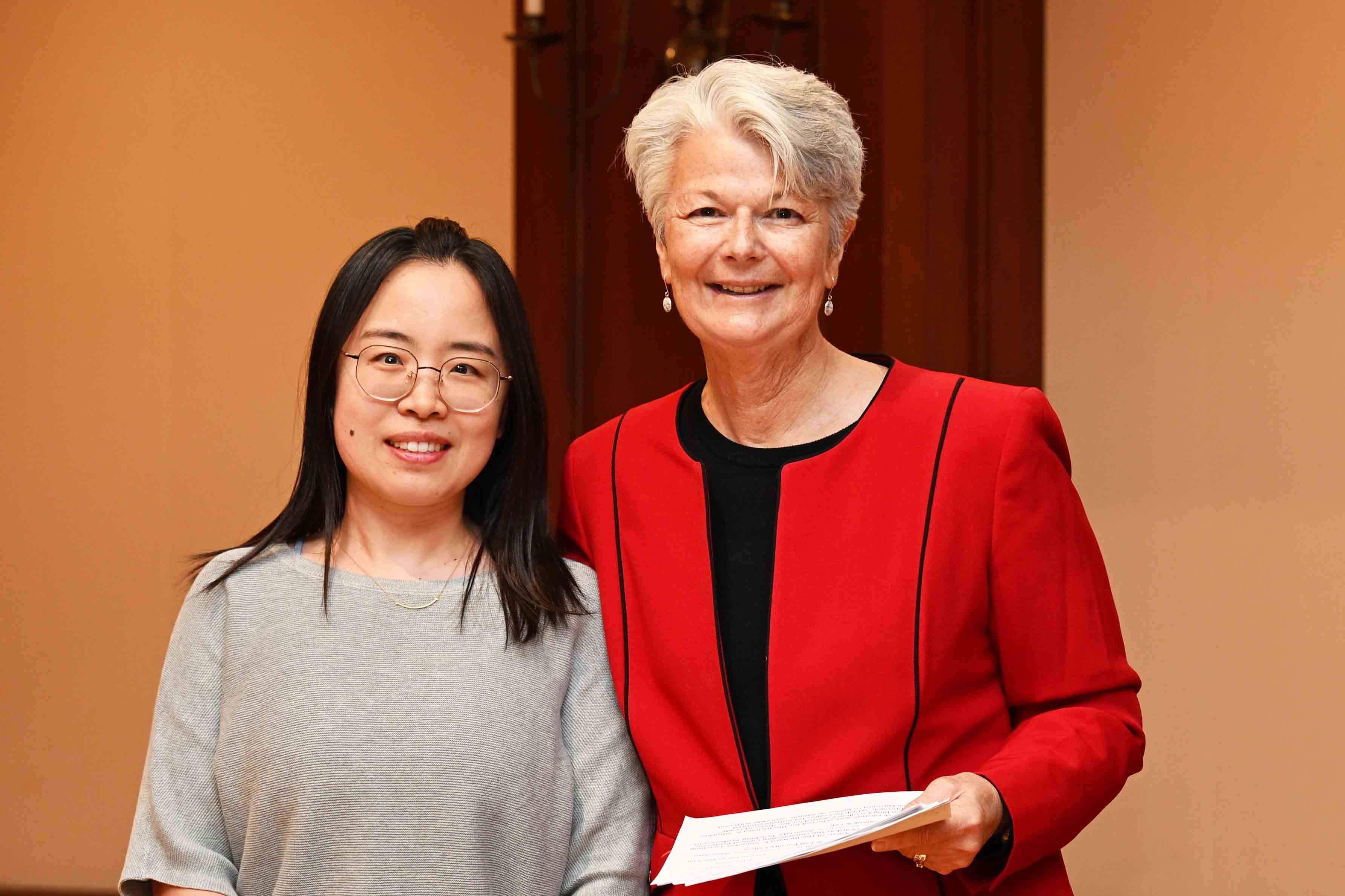 Two women pose together indoors, one wearing a red jacket and holding papers.