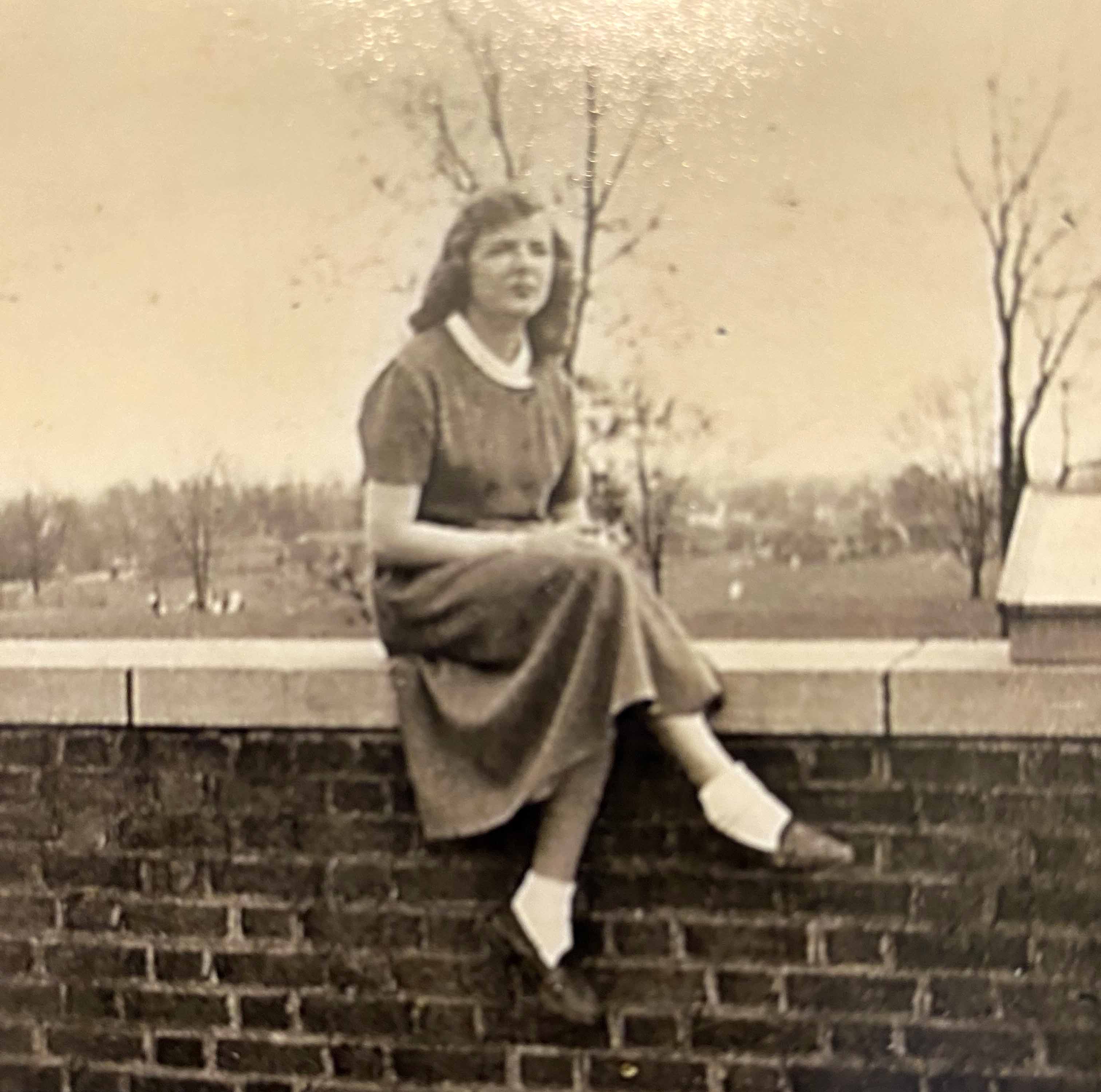 Vintage black-and-white photo of a young woman in a dress sitting on a brick wall, with bare trees and a distant landscape behind her.