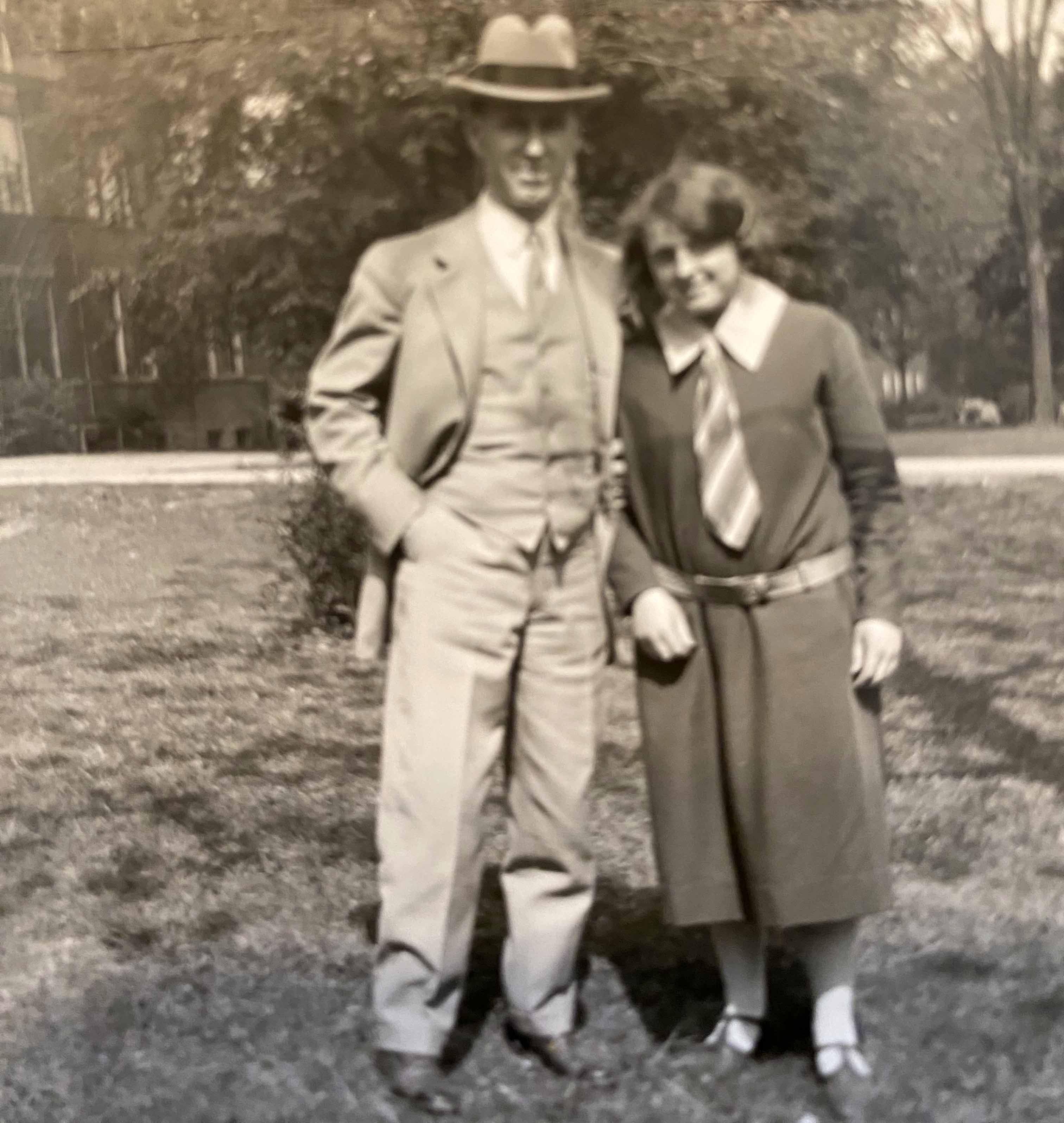 Vintage black-and-white photo of a couple standing outdoors on grass, with the man in a suit and hat and the woman in a dress with a wide collar.