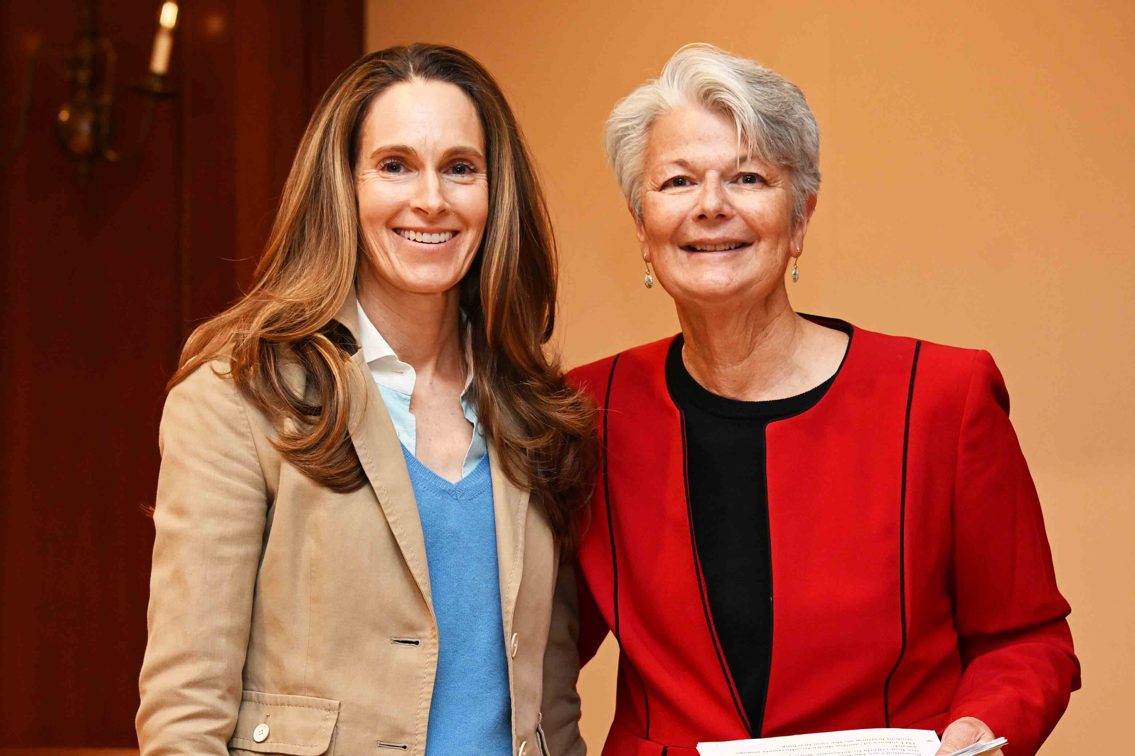 Two women smiling at an indoor event, one in a tan blazer and one in a red jacket holding papers.