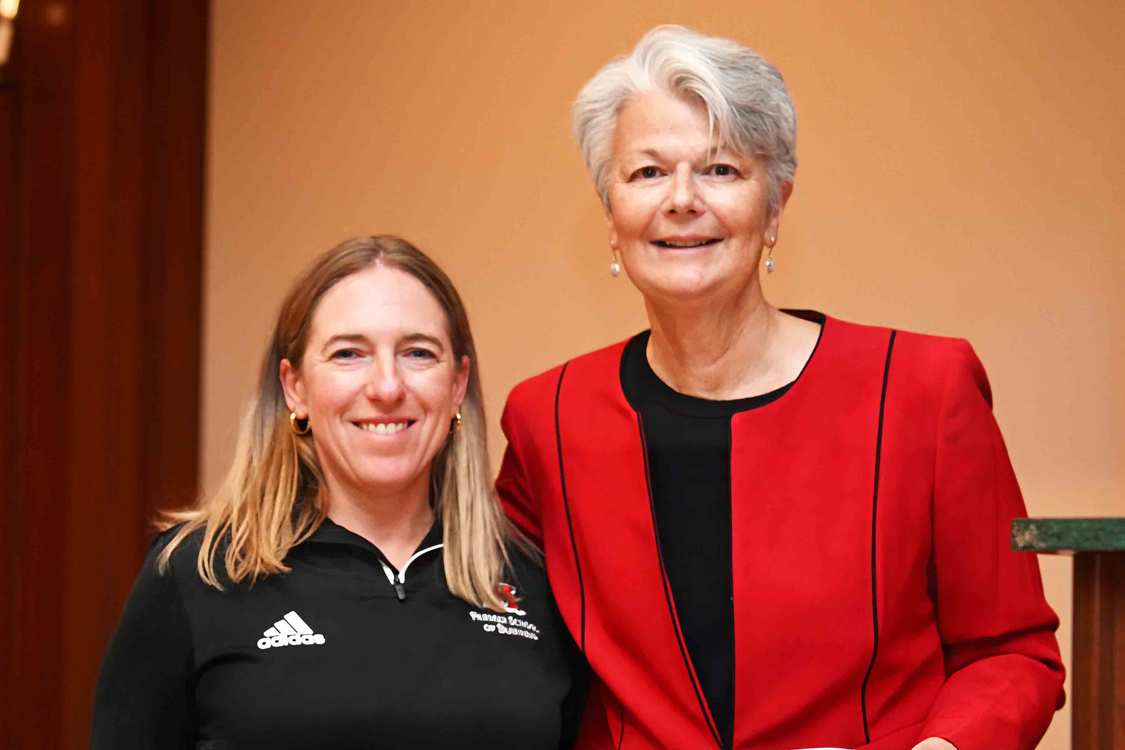 Two women smiling at an indoor event, one in a black athletic jacket and the other in a red blazer.