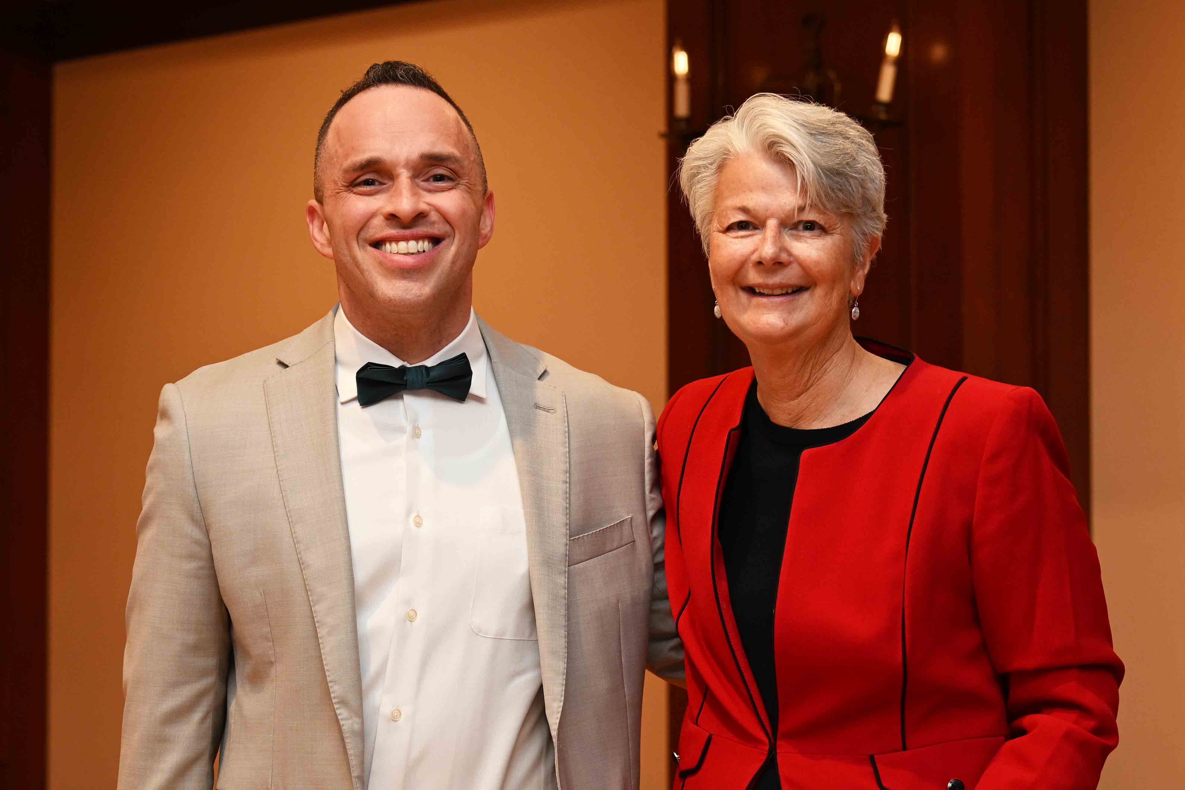 Two people smiling at a formal event, one in a light suit with a bow tie and the other in a red jacket.