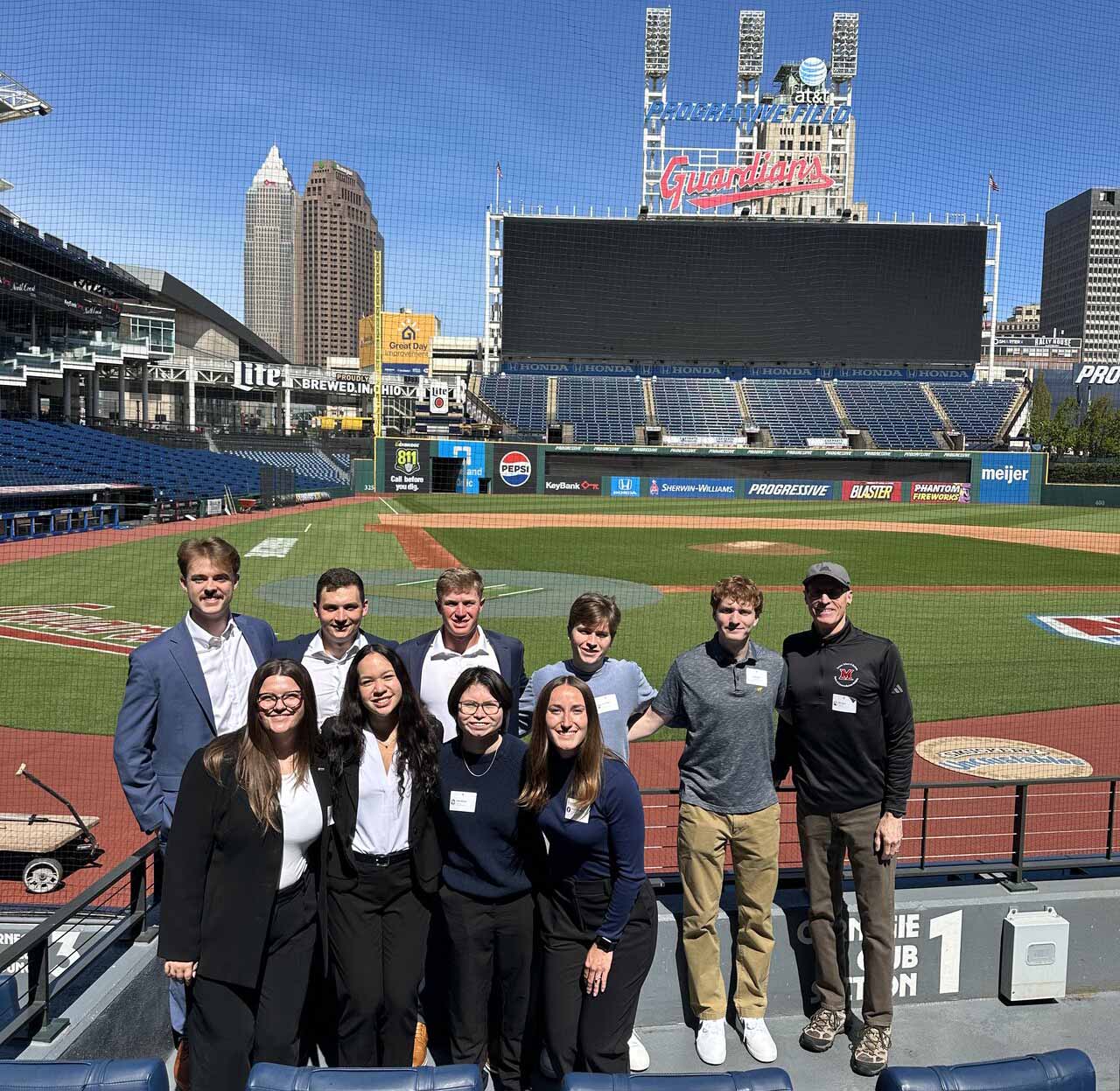 Group of nine people posing for a photo in the stands of a baseball stadium, with the field, scoreboard, and city skyline in the background.