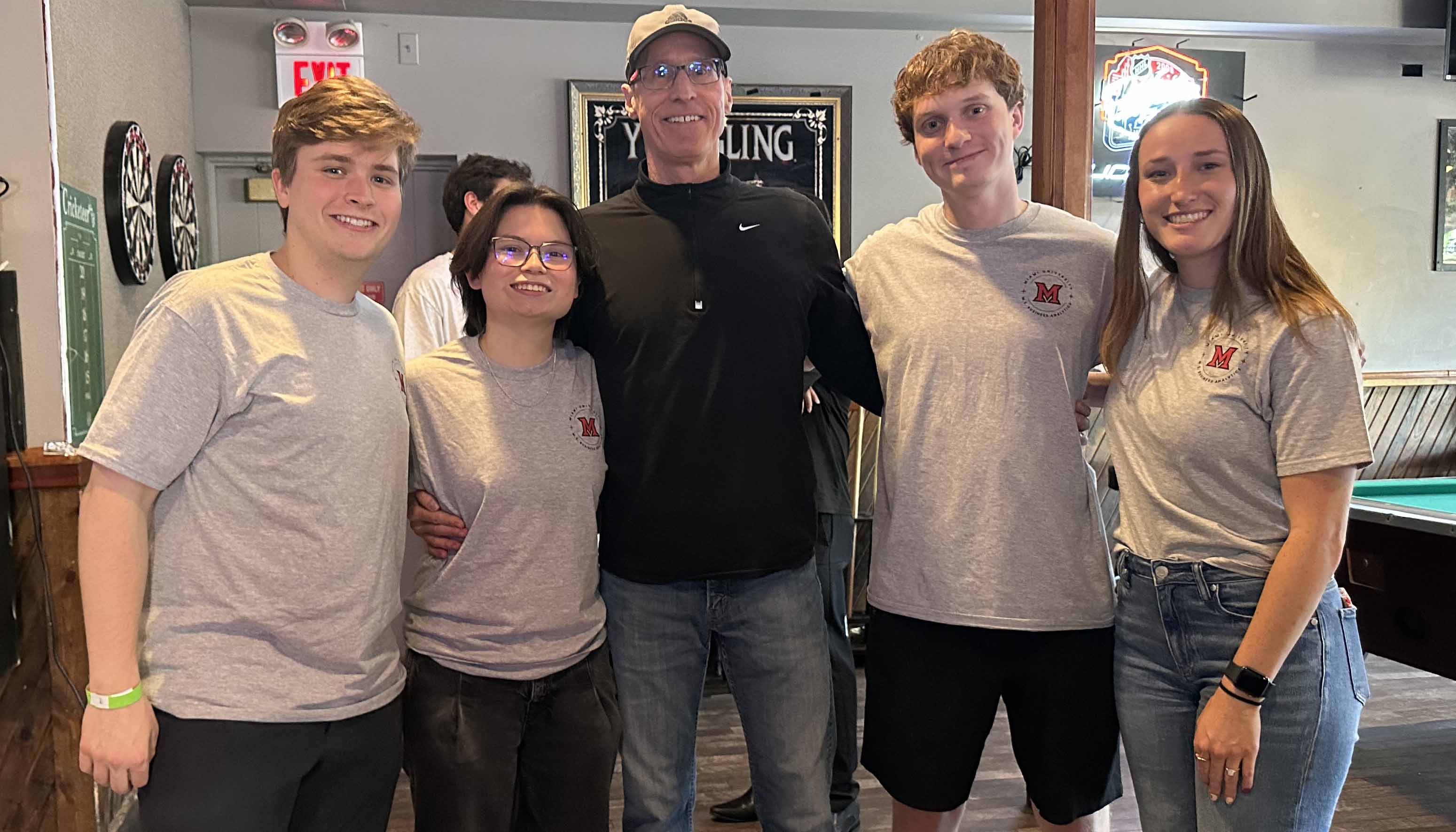 Group of five people smiling indoors, with four wearing matching gray shirts with a red “M” logo, standing near a dartboard and a pool table.