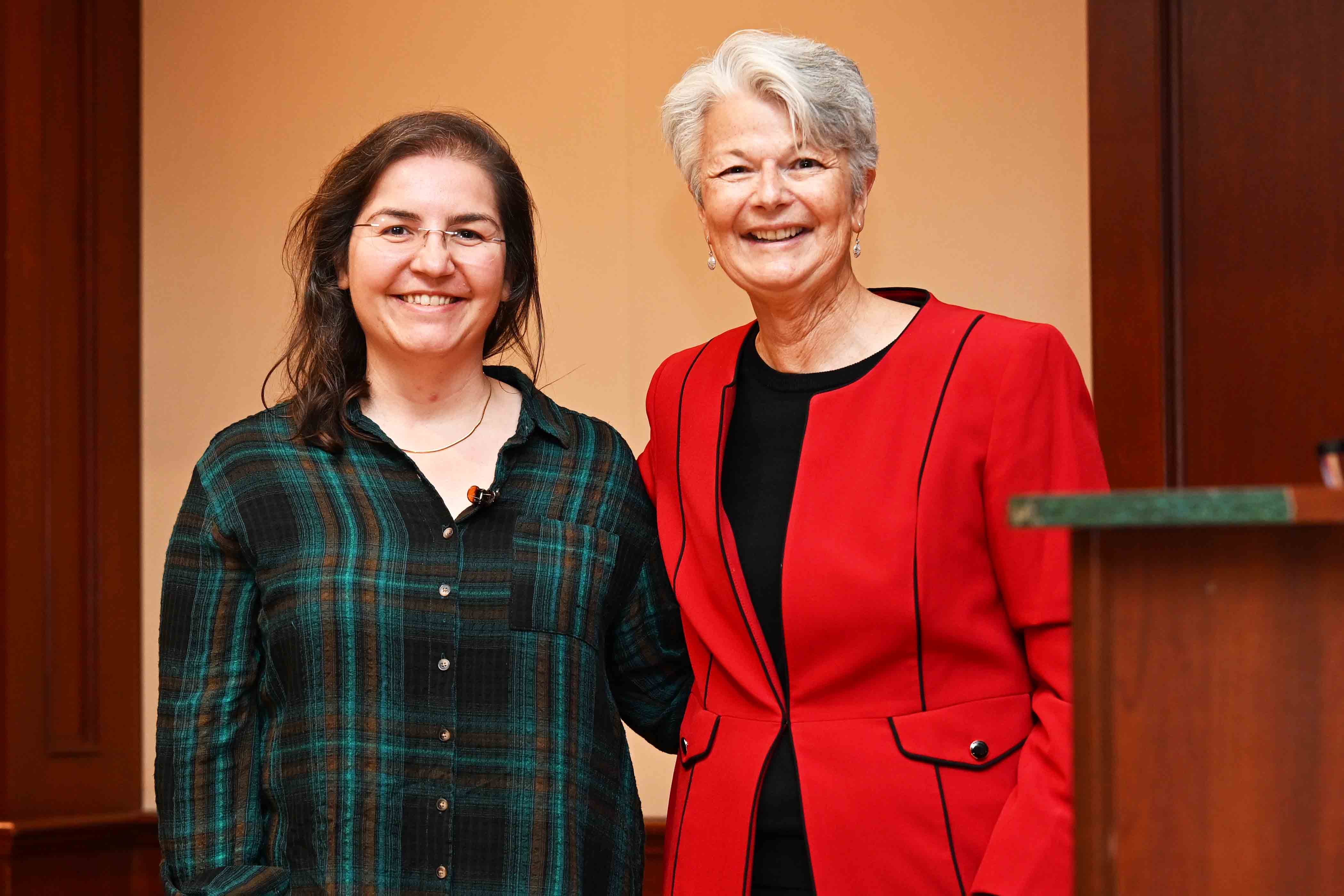 Two women smiling and posing together indoors, one wearing a plaid shirt and the other a red blazer.