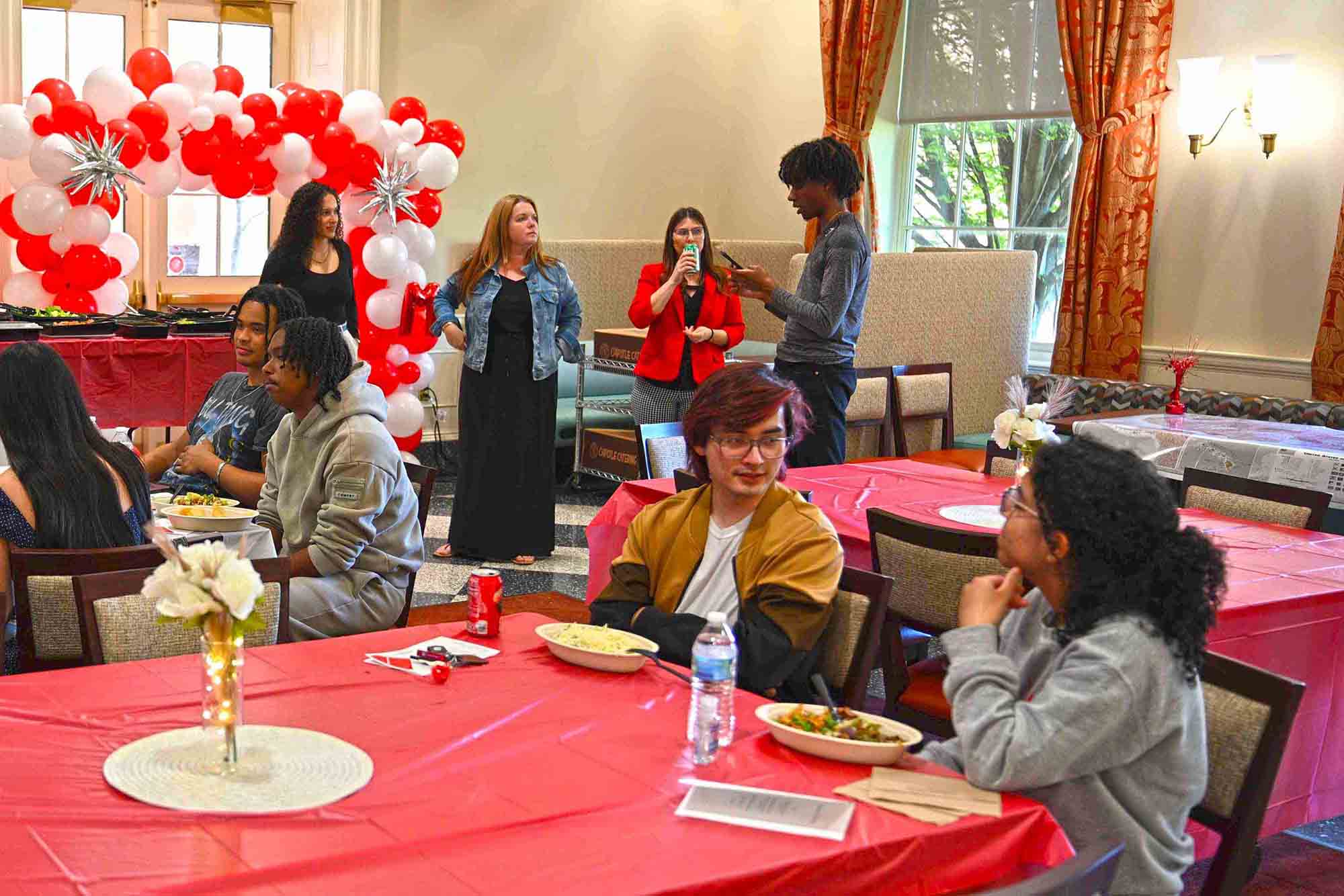 Group of people at a catered indoor event, seated at round tables with red tablecloths and a red-and-white balloon arch in the background.