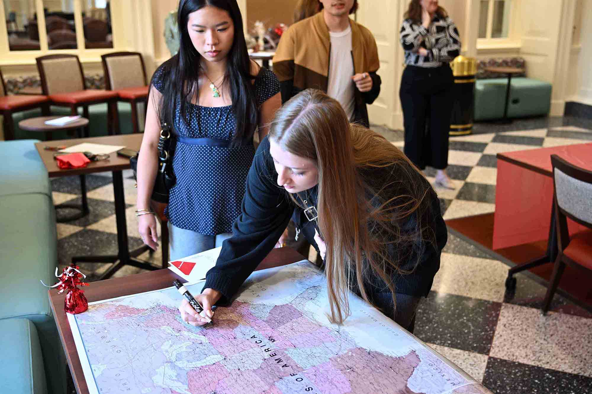 Two students lean over a large map on a table, with one writing on it using a marker while others stand nearby in a lounge area.