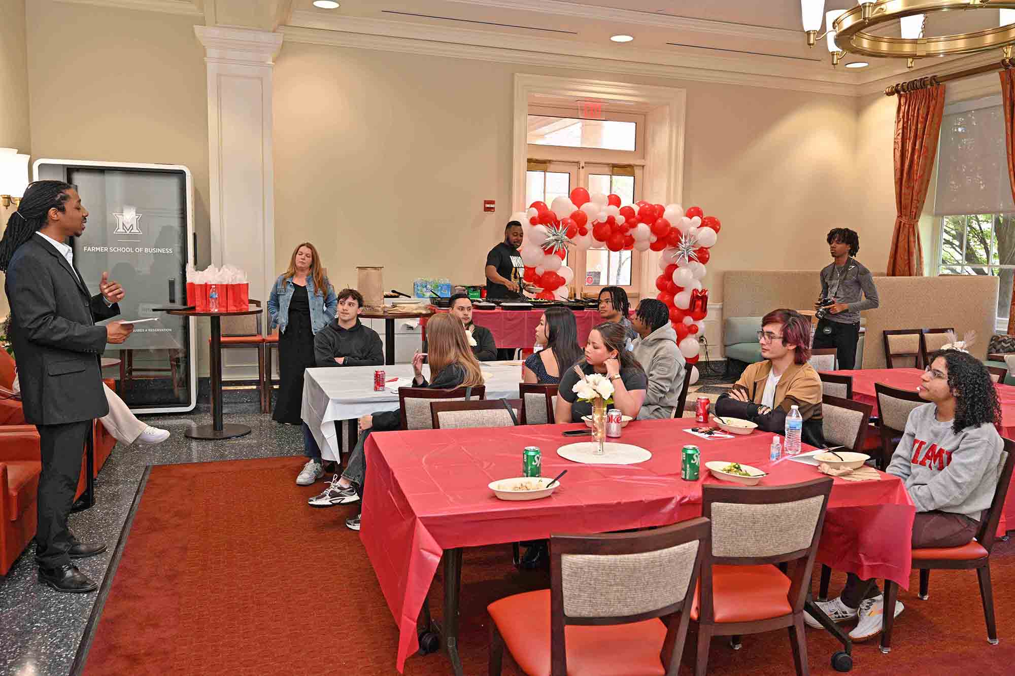 Speaker addresses a group seated at round tables during an indoor campus event, with red-and-white balloon decorations and a “Farmer School of Business” sign in the background.