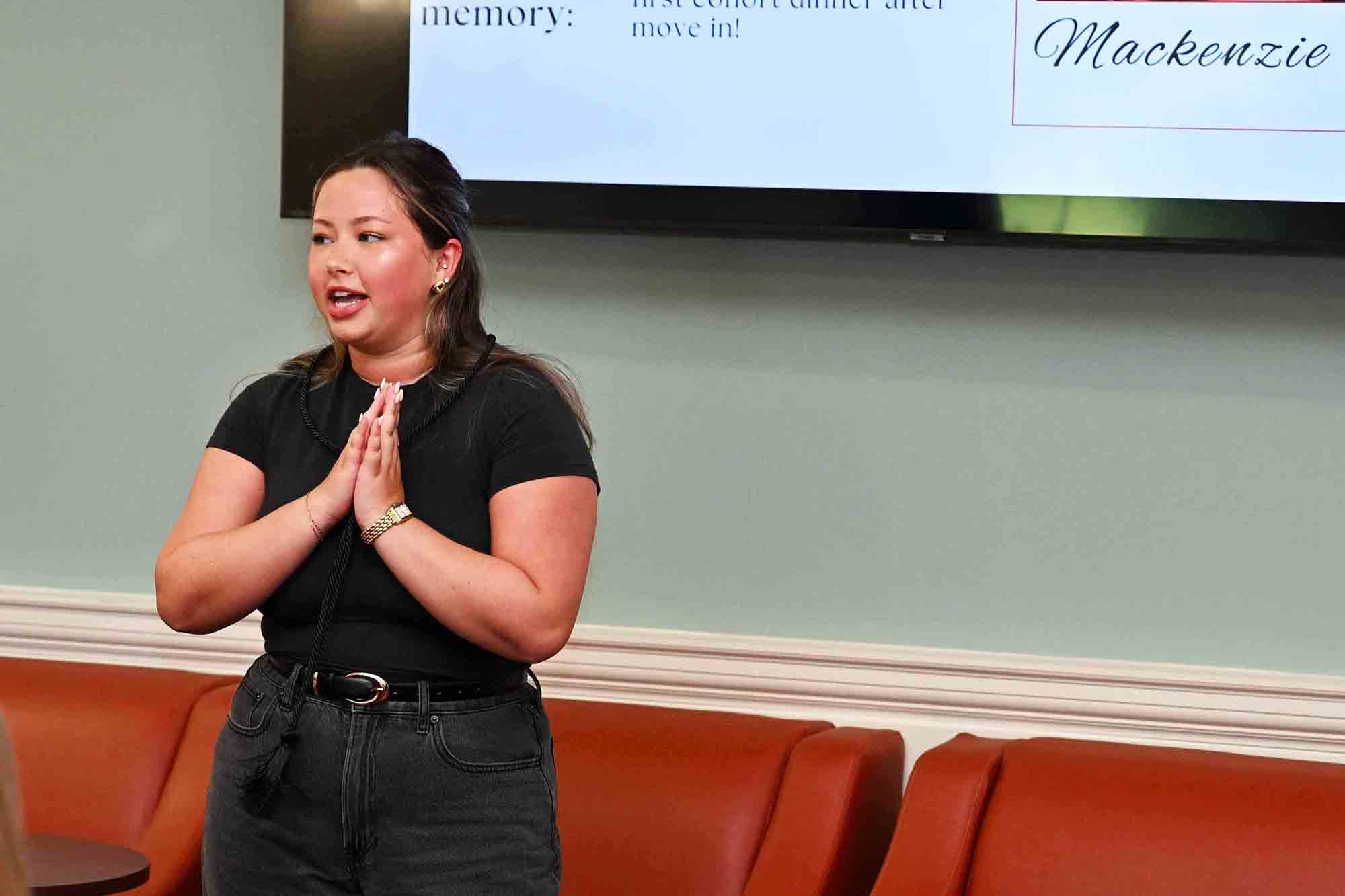 Woman speaking with hands clasped in front of a wall-mounted screen displaying a presentation slide.