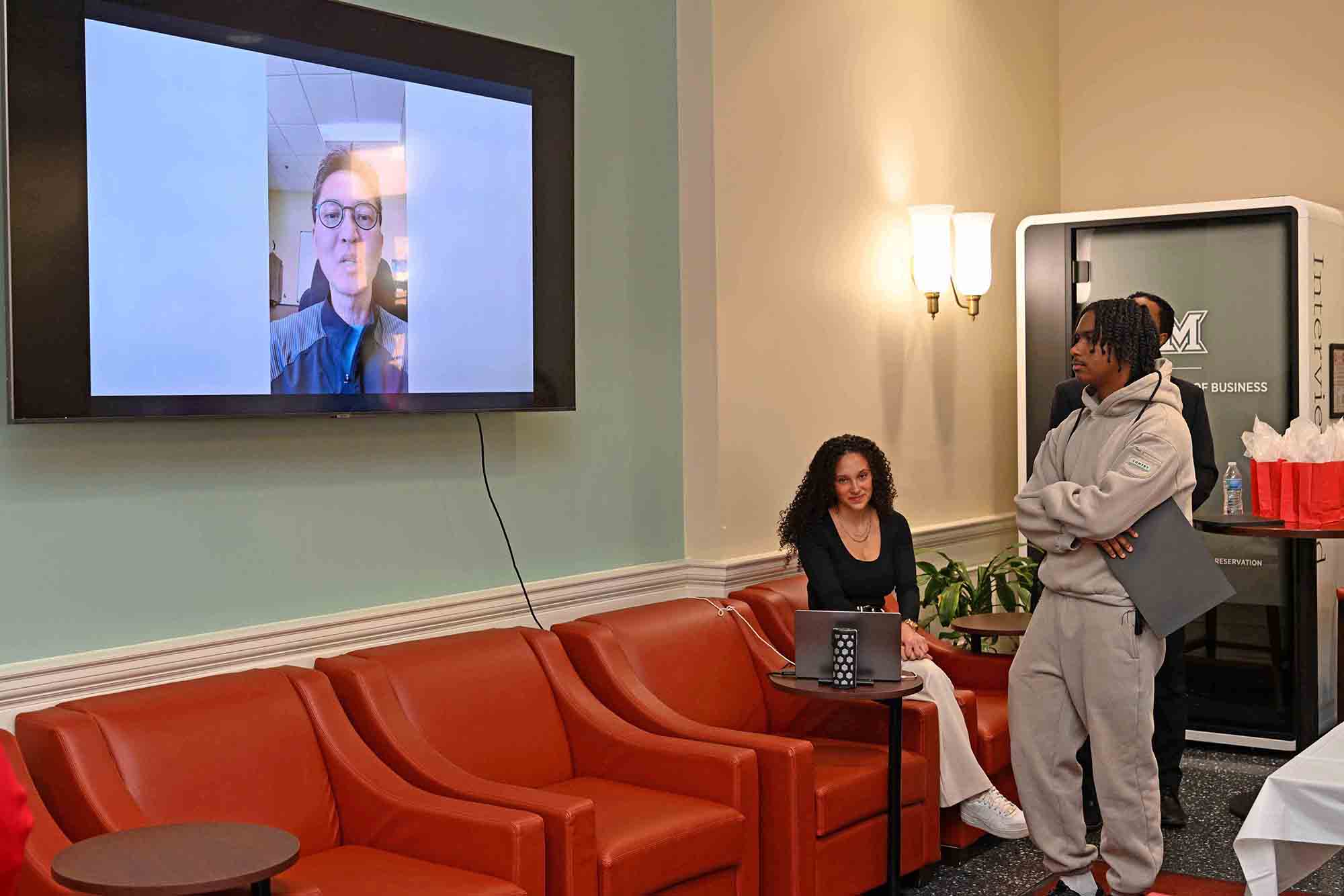 People in a lounge watch a video call displayed on a wall-mounted TV, while a student stands nearby holding a folder and another sits by a laptop.