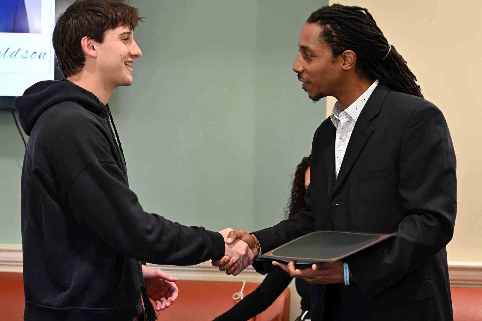 A man in a hoodie shakes hands with a man in a suit holding a folder, as if congratulating him at a presentation or ceremony.