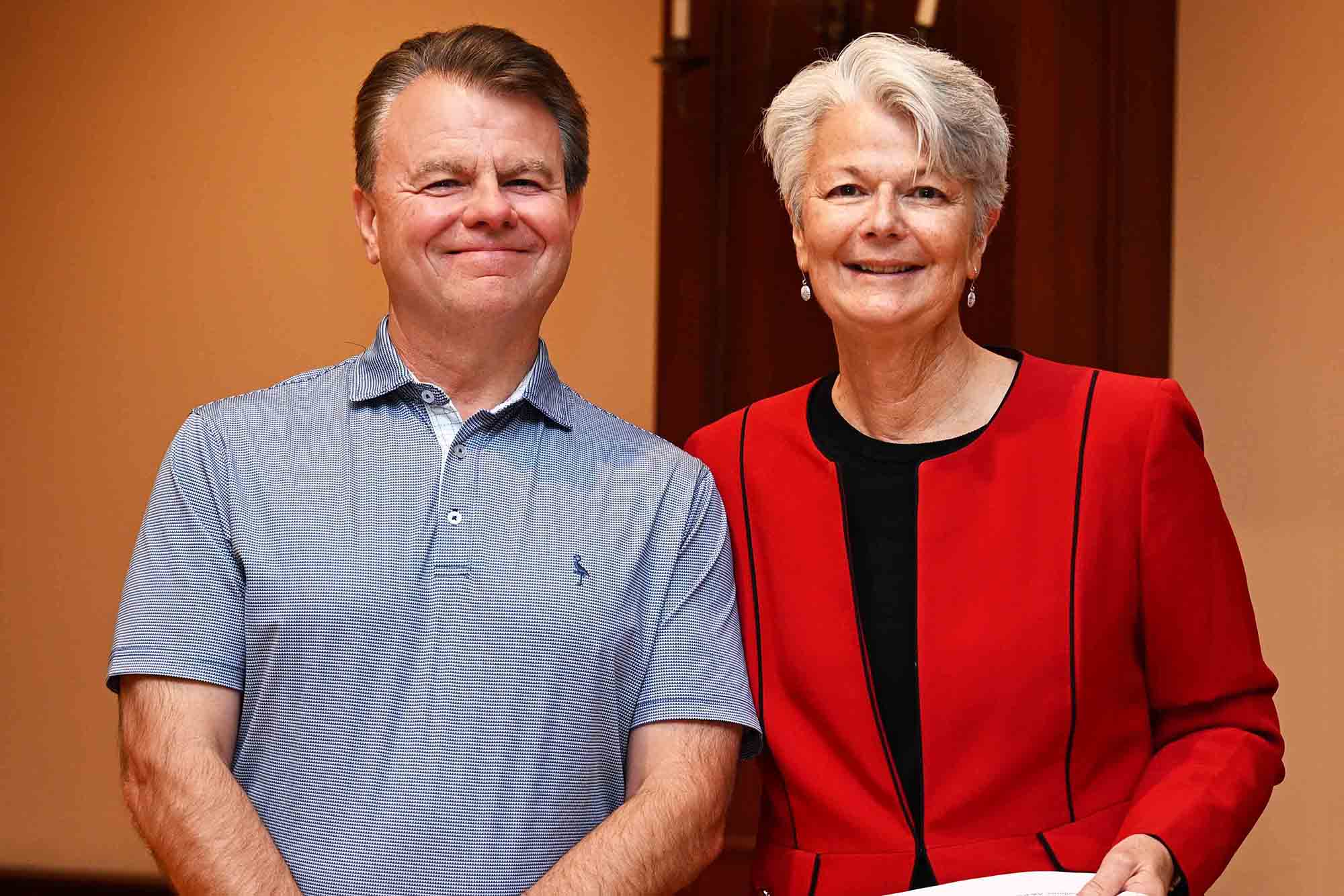 Two people smiling indoors, standing side by side, with one wearing a red blazer and the other a blue polo shirt.