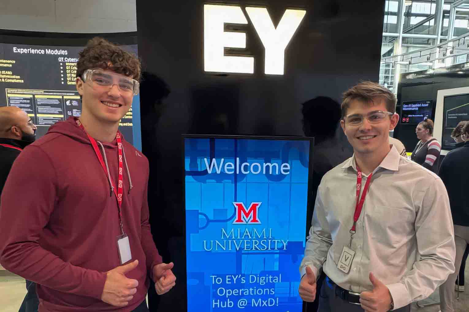 Two people wearing safety glasses give thumbs up beside a screen that says “Welcome Miami University” at an EY booth.