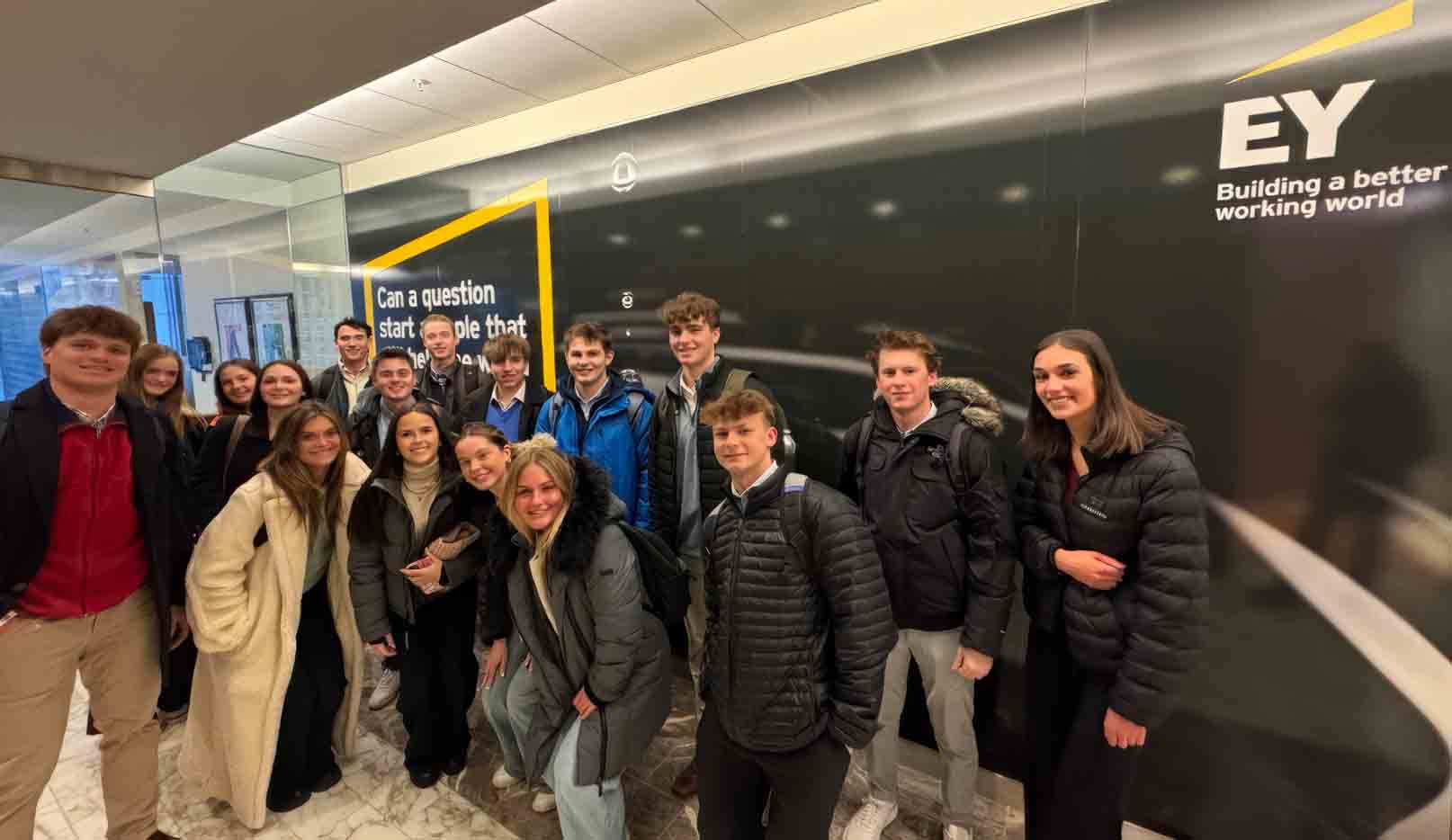 Group of students posing in an office lobby beside a wall mural featuring the EY logo and the slogan “Building a better working world.”